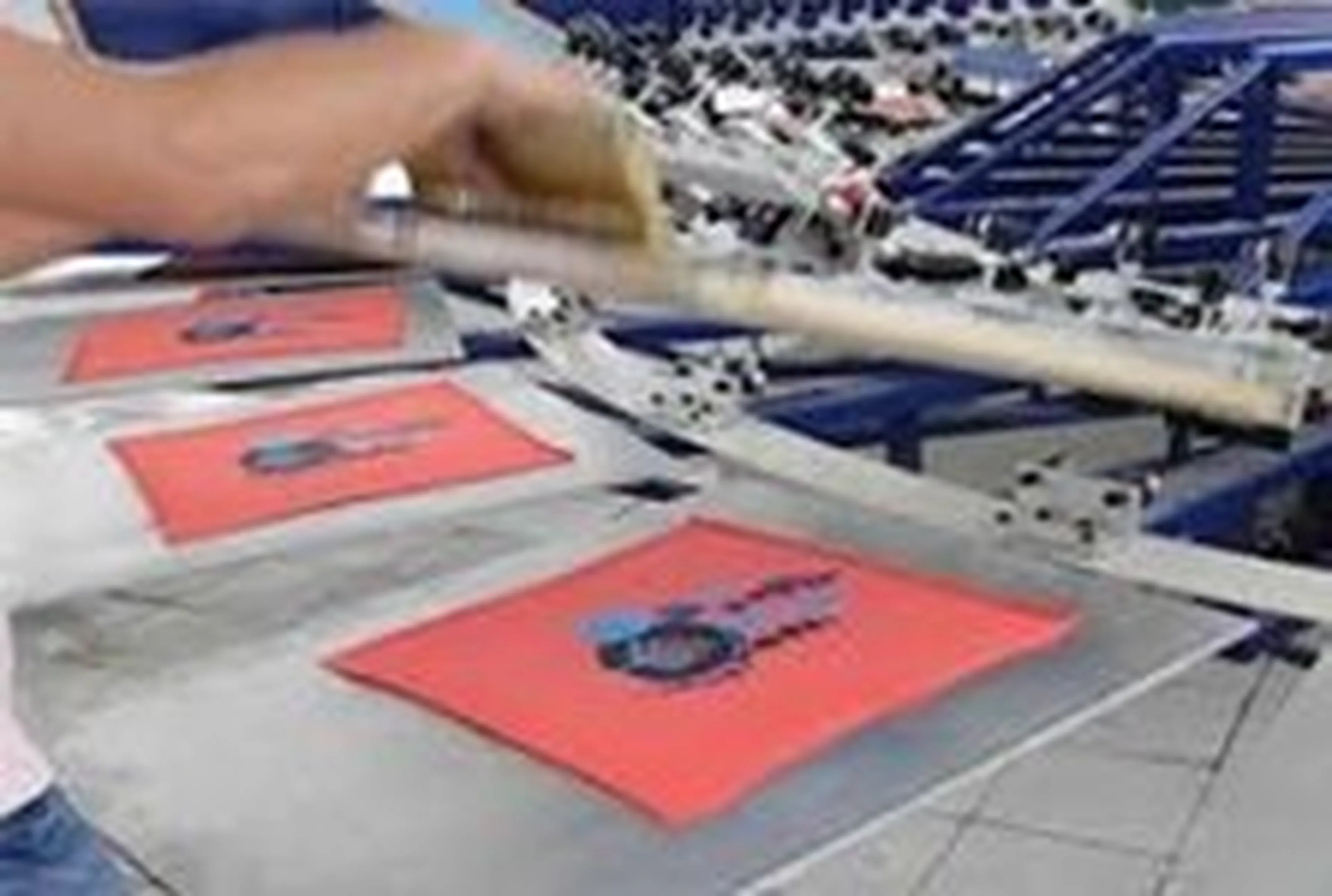 A person operates a screen printing machine, transferring designs onto red fabric.