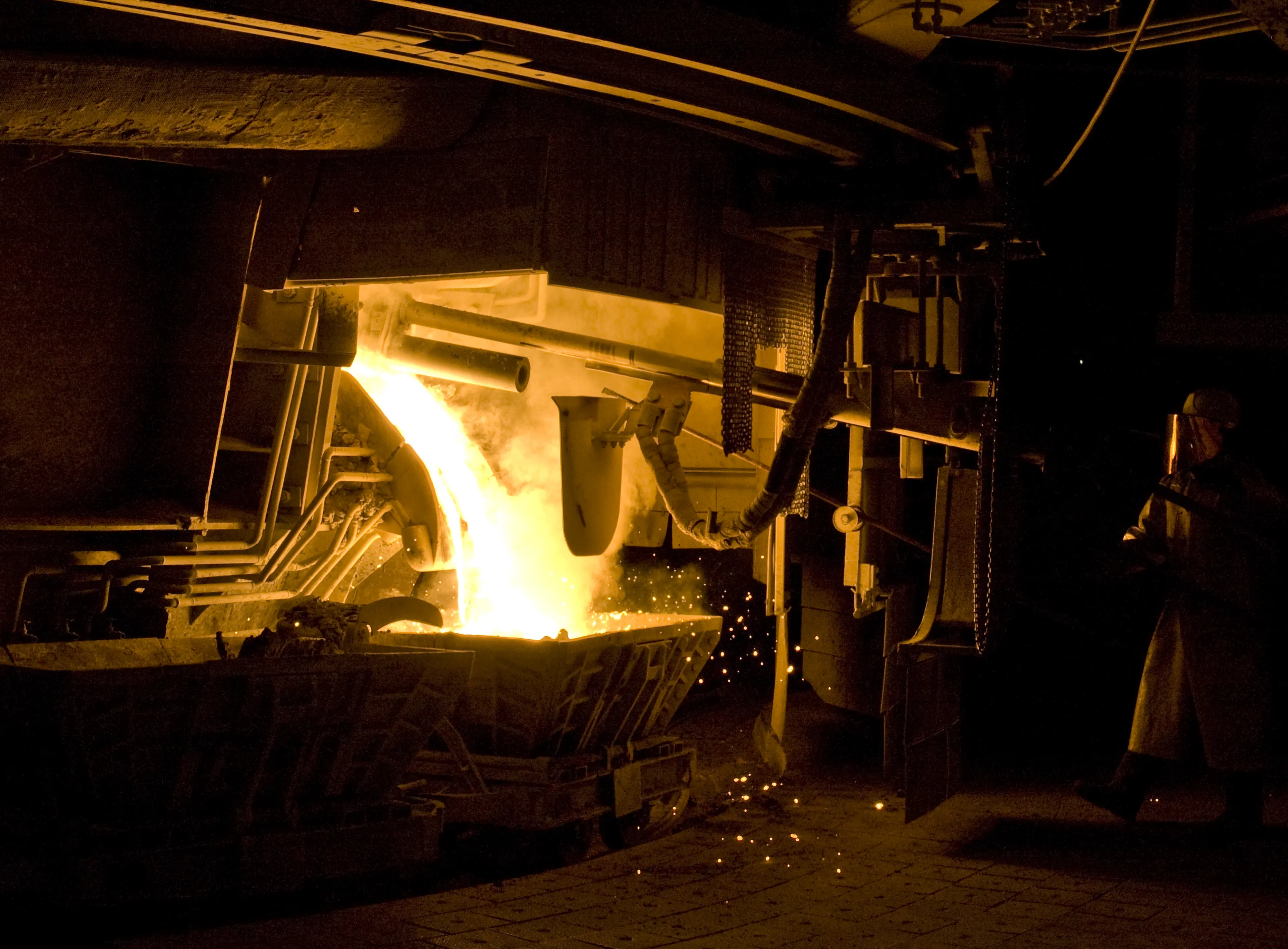 Molten metal pours from a furnace into a container, illuminating a dark industrial setting with a worker in protective gear nearby.