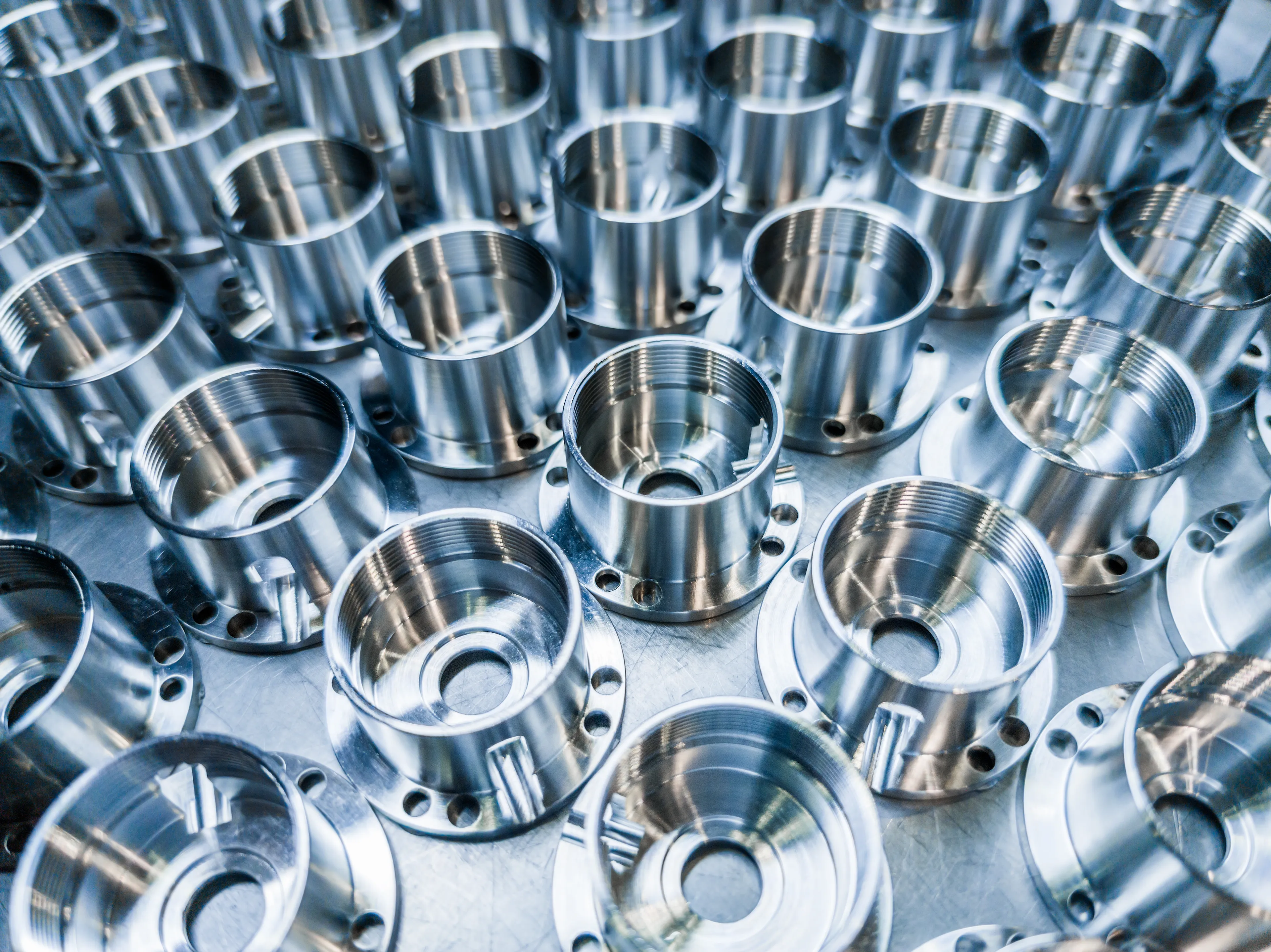 Rows of shiny metallic cylindrical components with threaded openings and multiple drilled holes, arranged on a reflective surface.