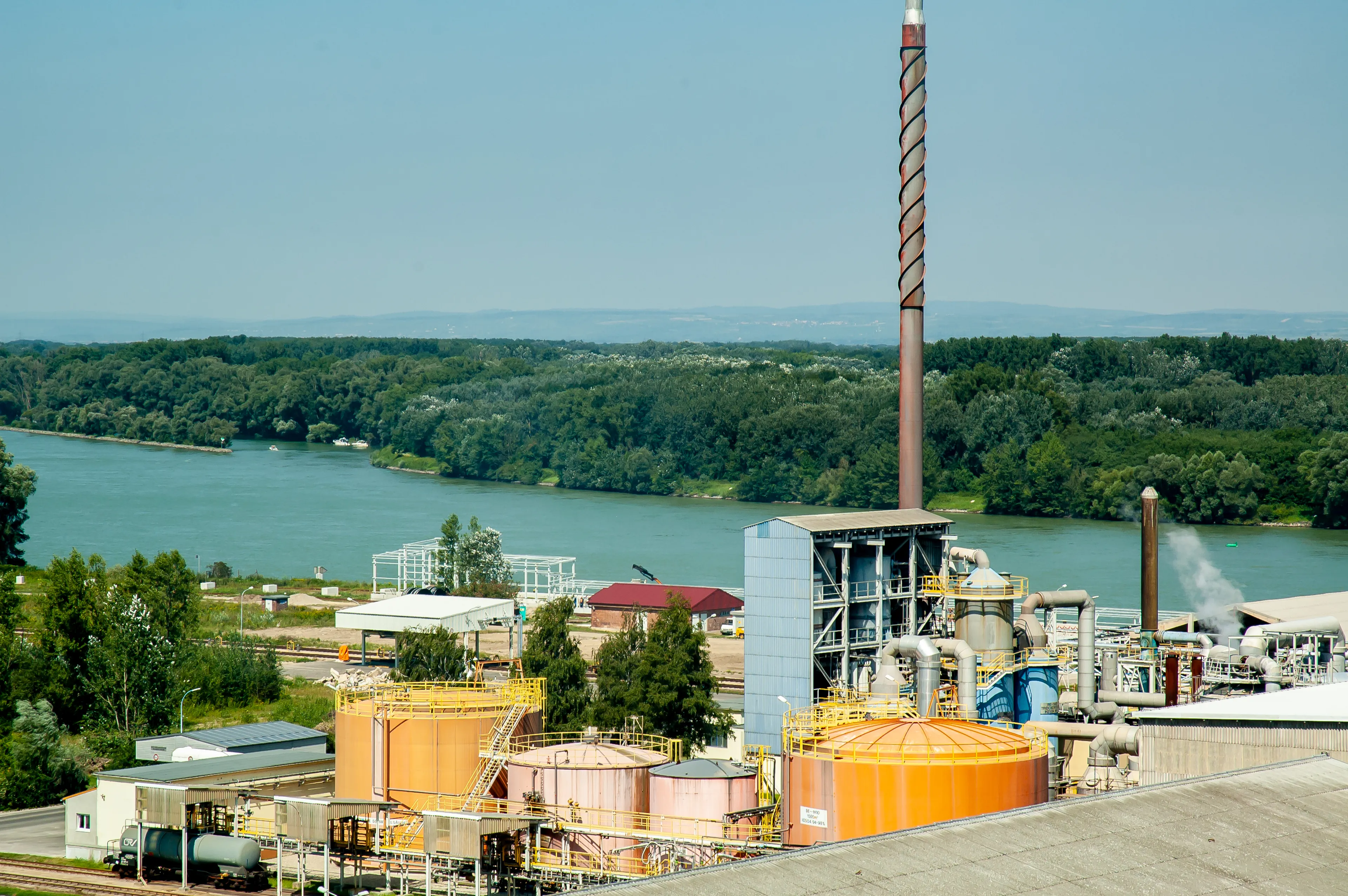 Industrial facility with large tanks and chimneys near a river, surrounded by trees and greenery in the background under a clear sky.