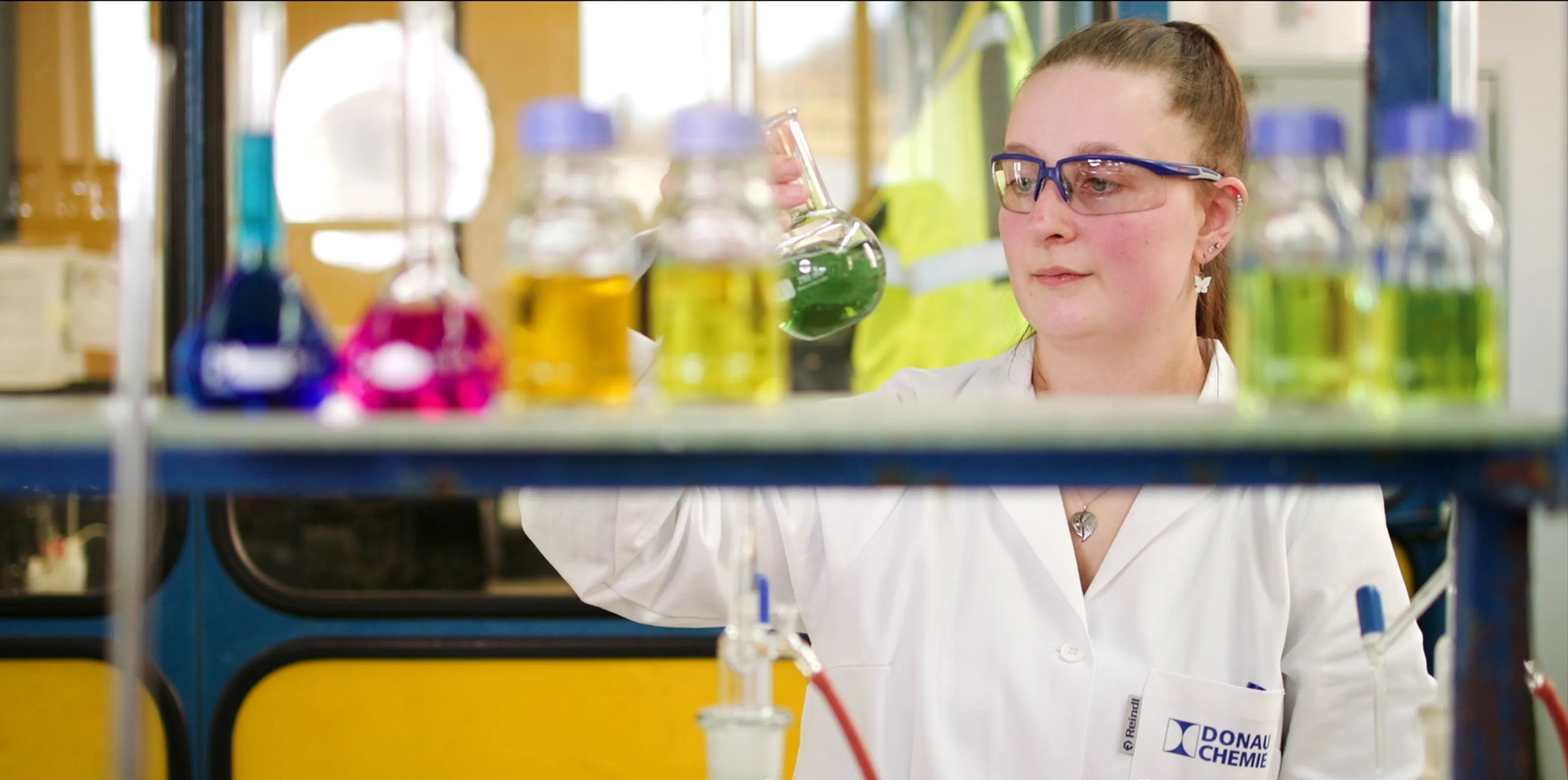 A chemist in a lab coat examines a green liquid in a flask, surrounded by colorful chemicals in bottles on a shelf.