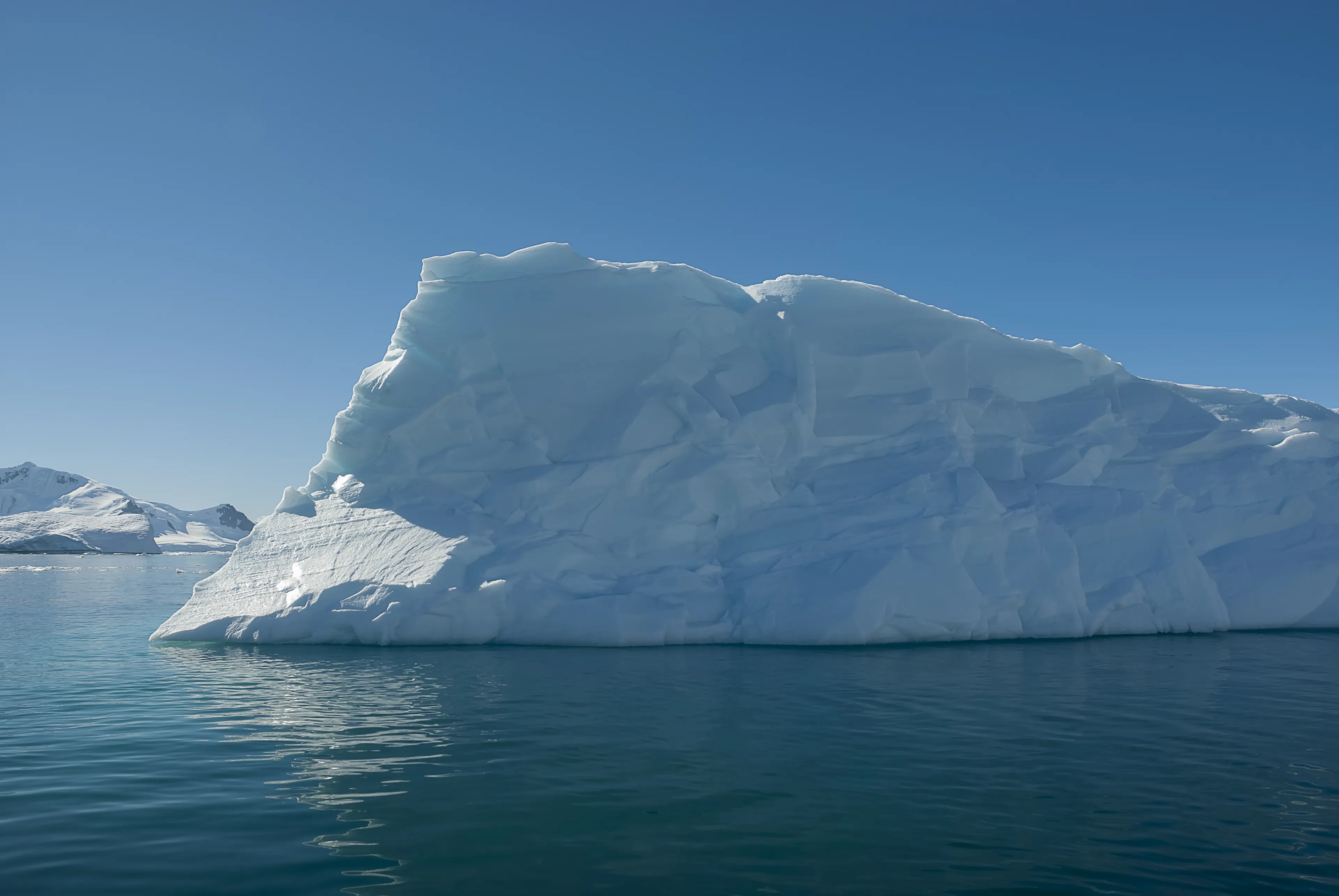 Ein großer Eisberg, der in ruhigem, blauem Wasser unter klarem Himmel treibt, mit verschneiten Landschaften im Hintergrund.