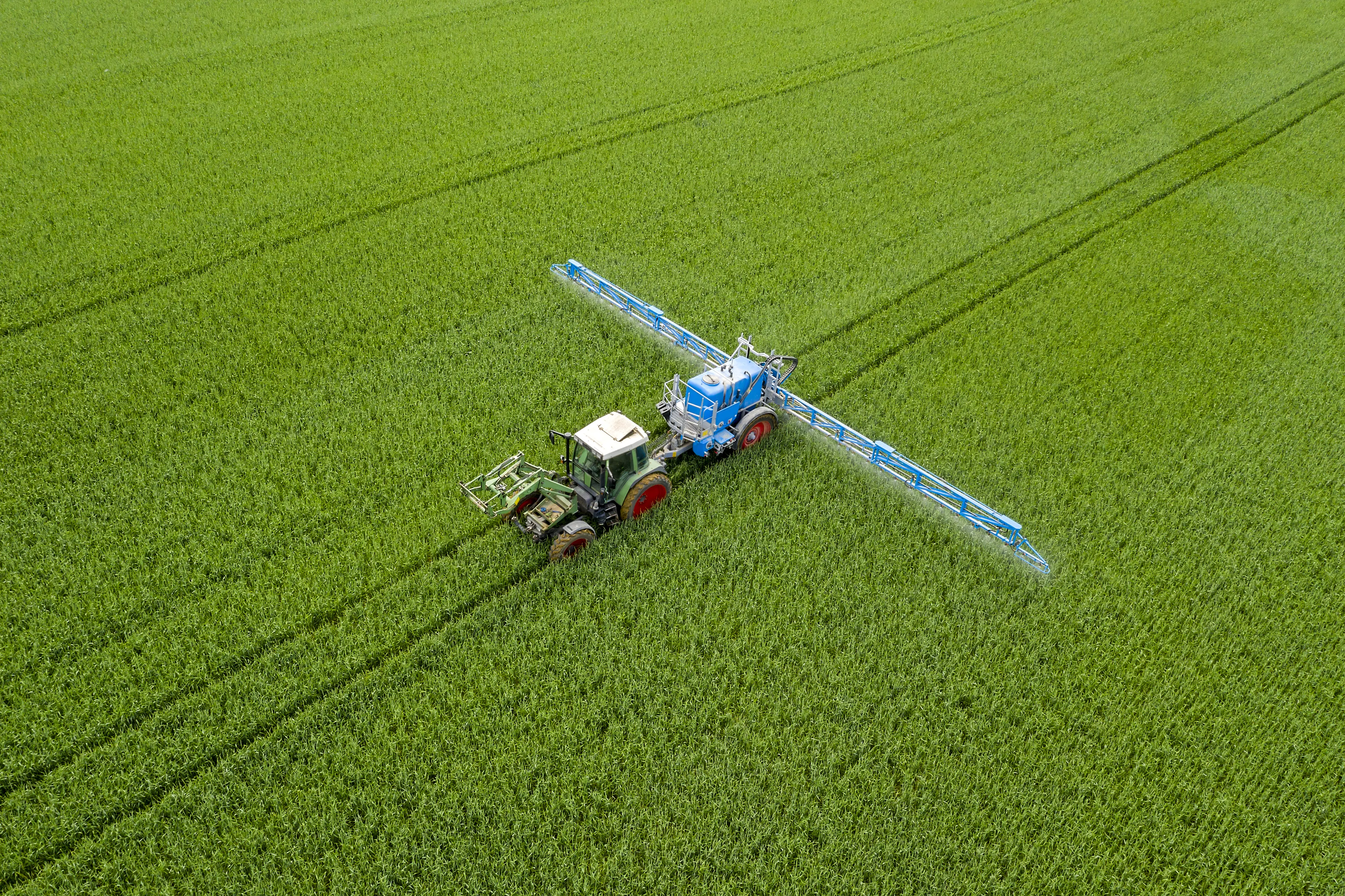 Aerial view of a tractor spraying pesticides over a vast green field, creating symmetrical lines through the crops.
