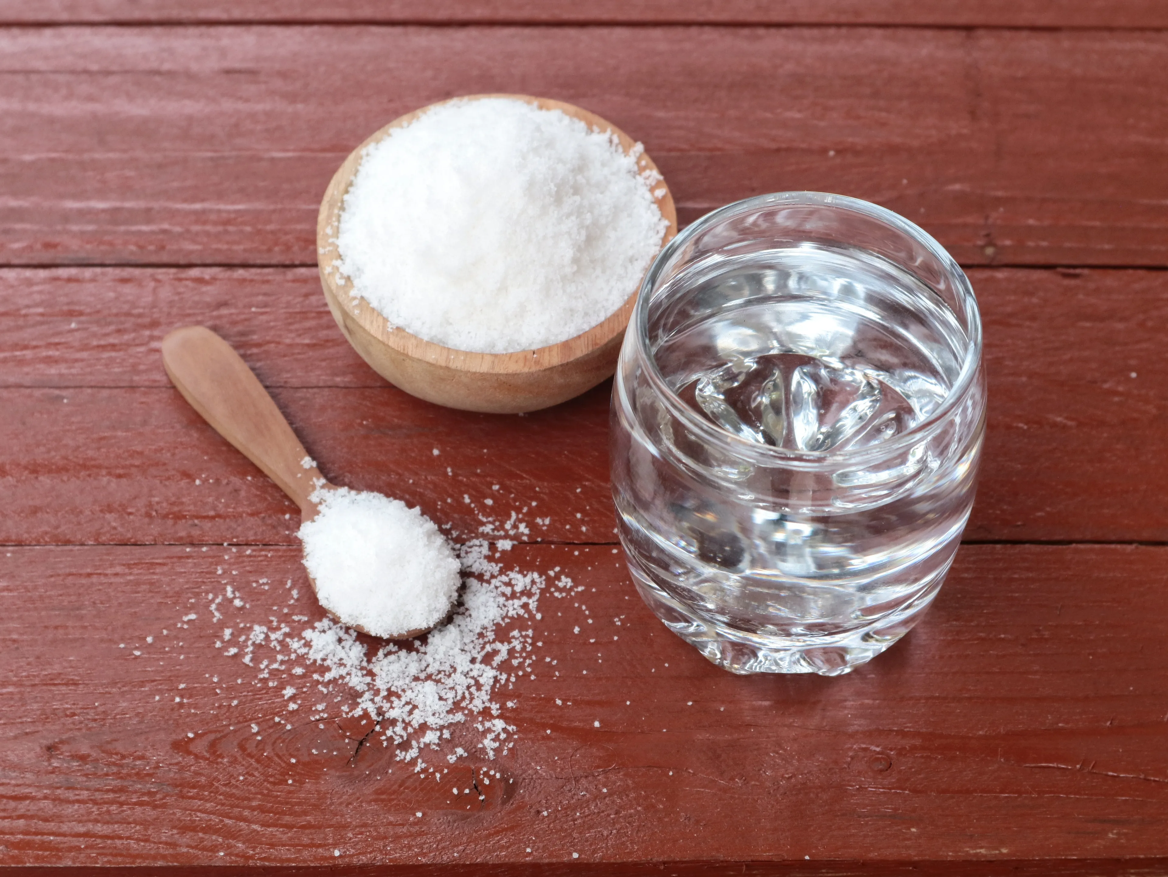A wooden bowl and spoon with salt and a glass of water are on a brown wooden table.