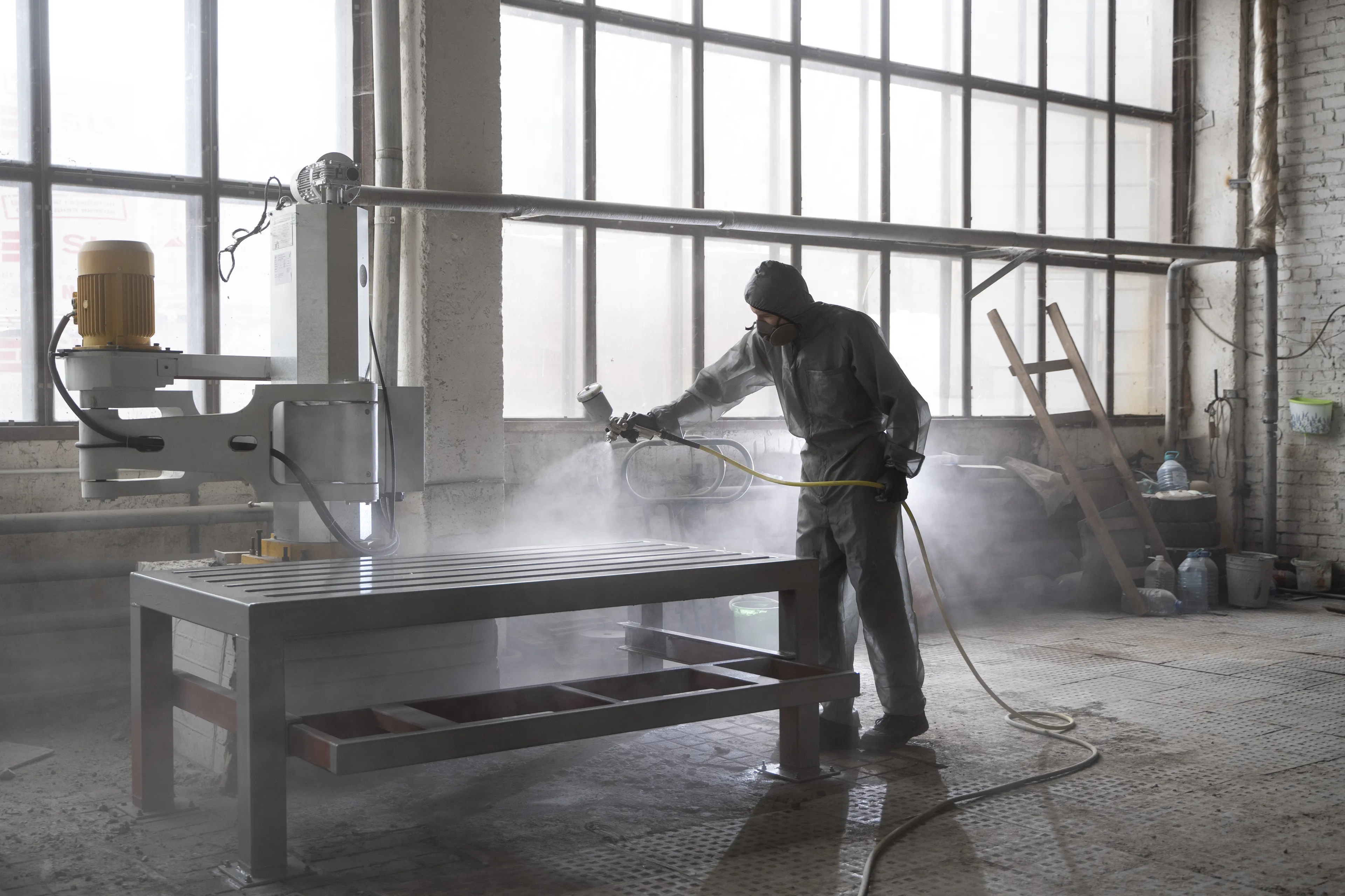 A worker in protective gear operates a spray gun on a metal table in a spacious industrial workshop with large windows.