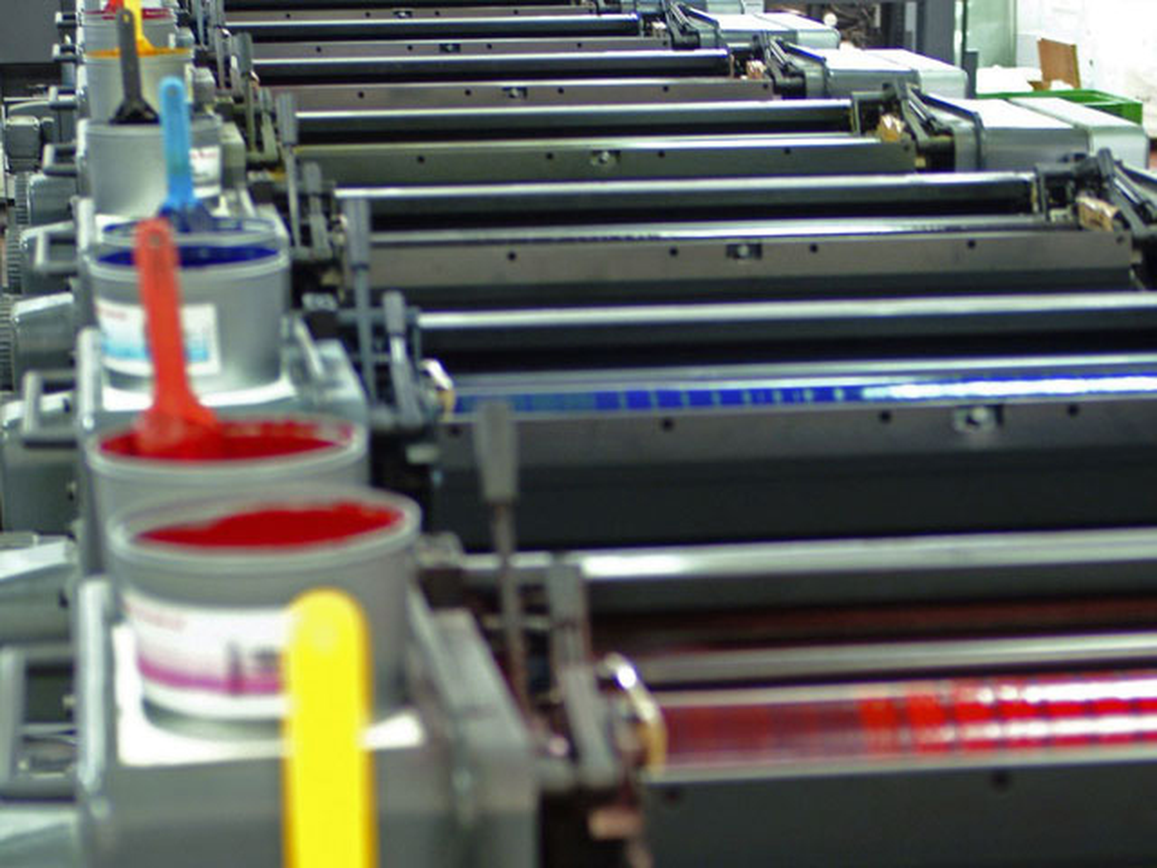 Close-up of an offset printing machine with colorful ink cans and rollers aligned in sequence, showing vibrant red, blue, yellow, and cyan.
