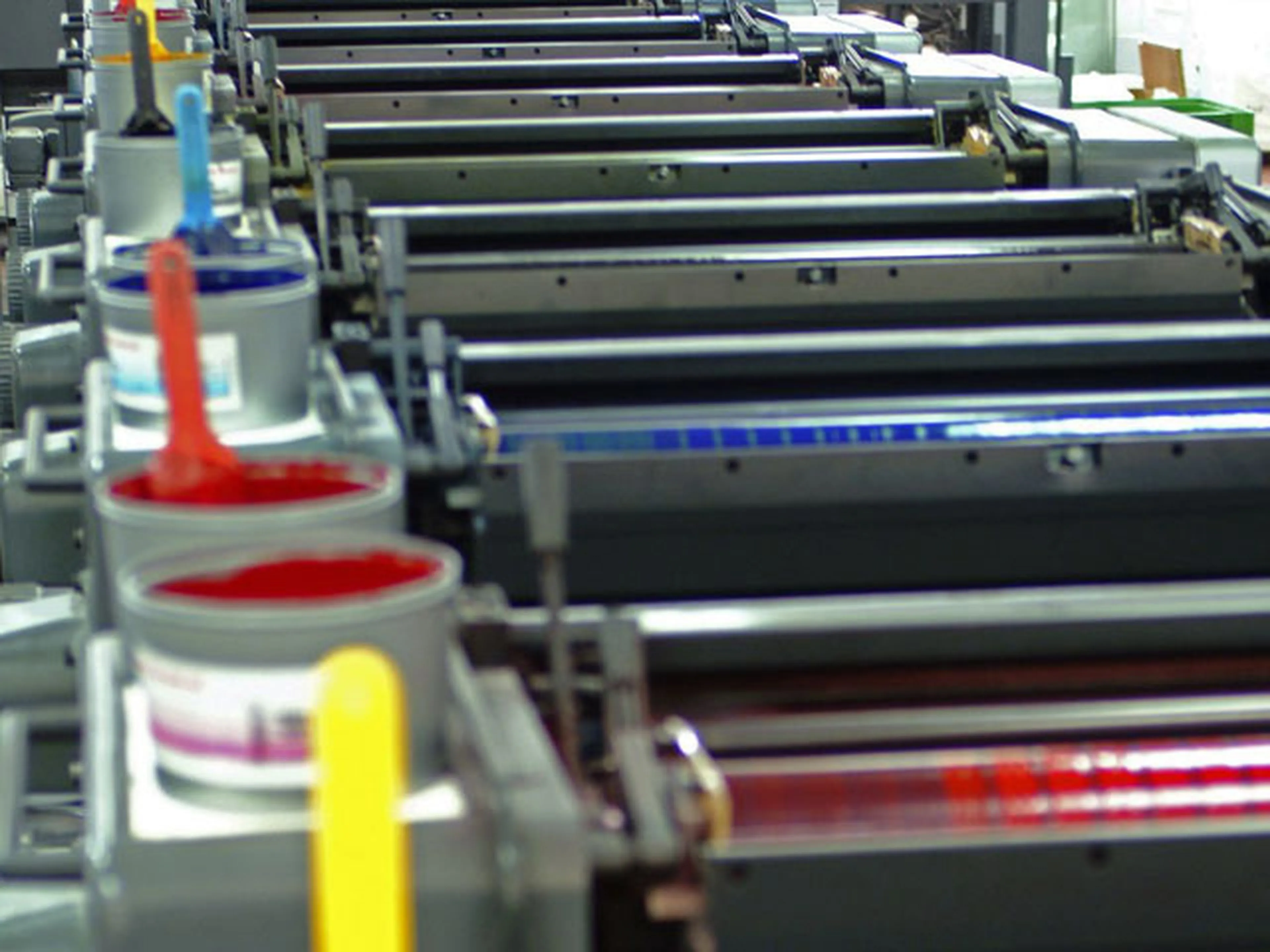 Close-up of an offset printing machine with colorful ink cans and rollers aligned in sequence, showing vibrant red, blue, yellow, and cyan.