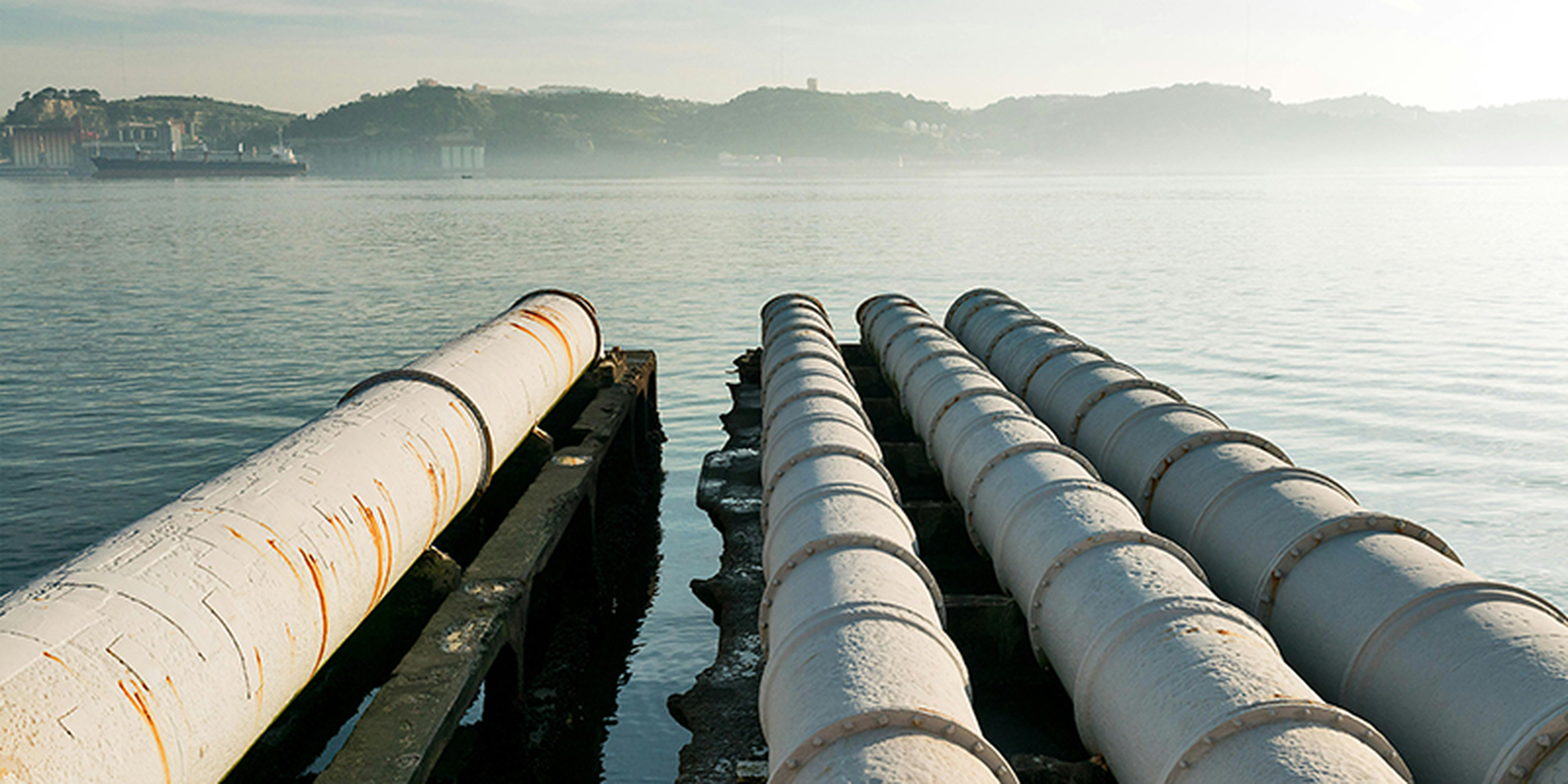 Large industrial pipes extend over calm water, with misty hills and a ship in the background under a hazy sky.