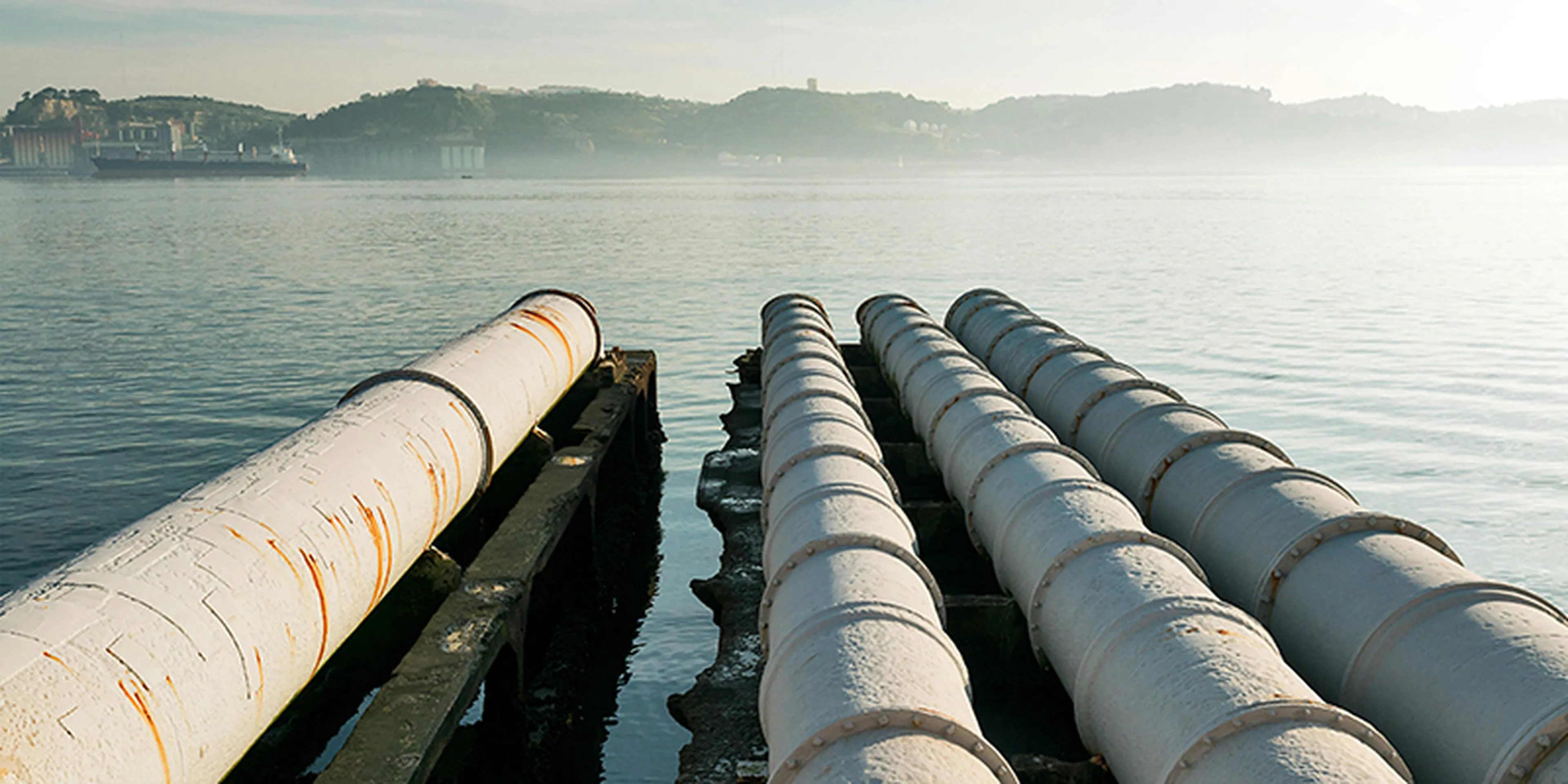 Large industrial pipes extend over calm water, with misty hills and a ship in the background under a hazy sky.