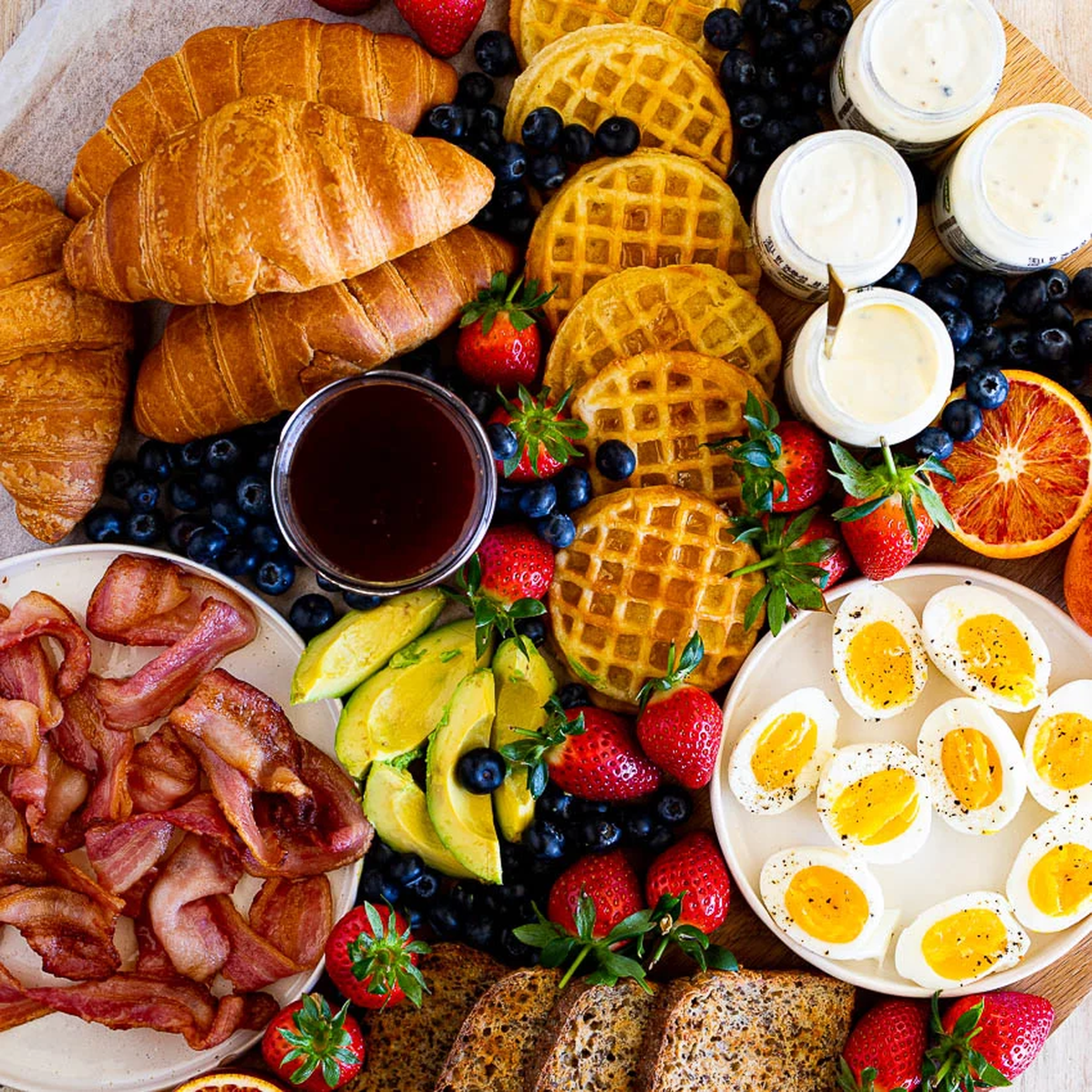 A breakfast spread with croissants, waffles, bacon, boiled eggs, avocado, berries, toast, yogurt, and syrup on a wooden surface.