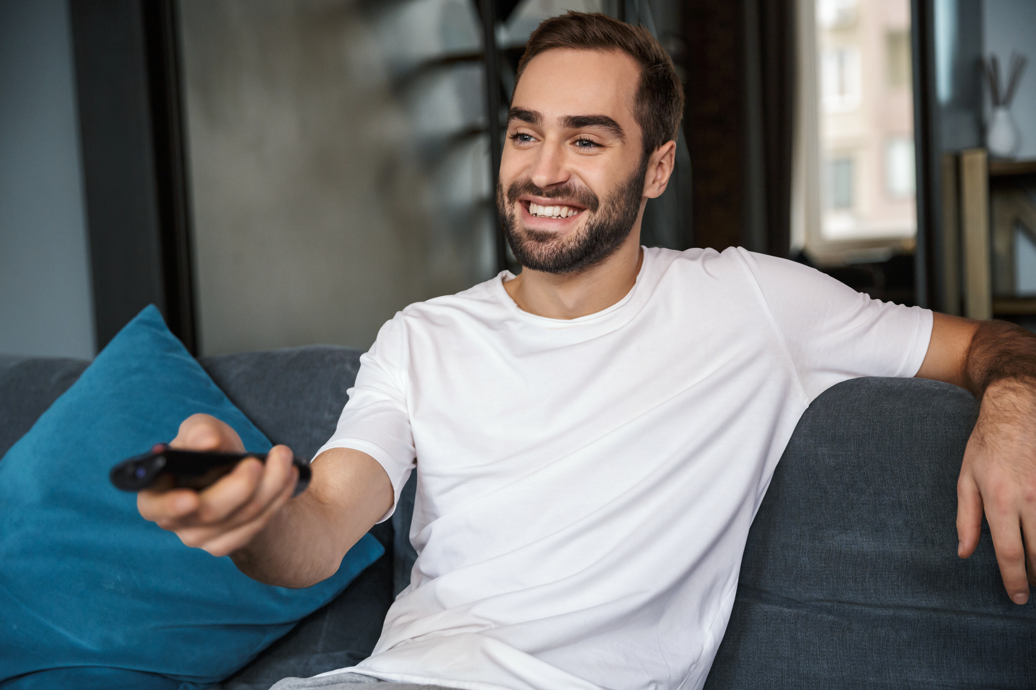Smiling man in a white t-shirt sits on a sofa, holding a TV remote, with a blue cushion beside him.