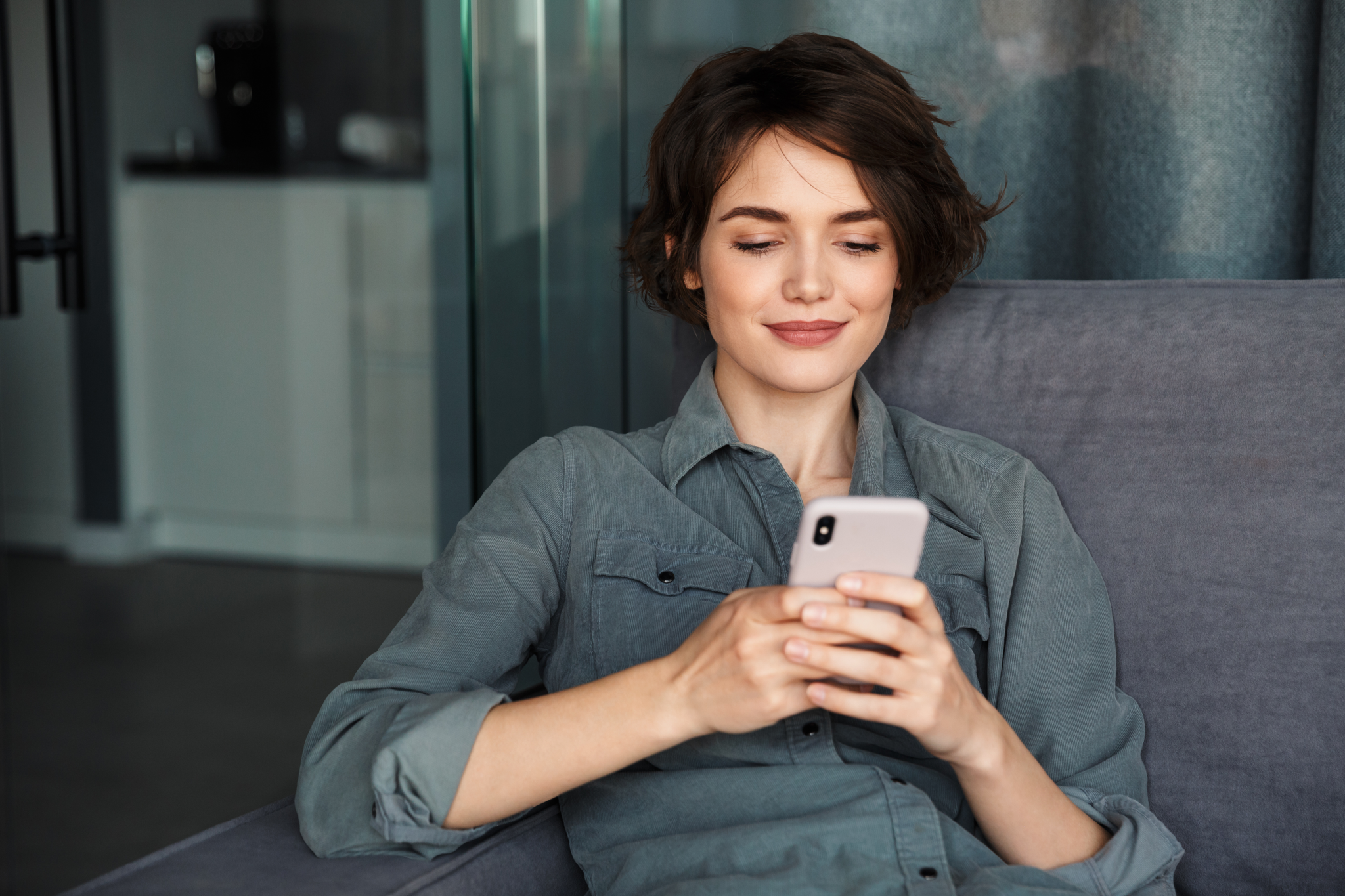 Woman with short hair, wearing a gray shirt, sits on a couch smiling while looking at her smartphone.