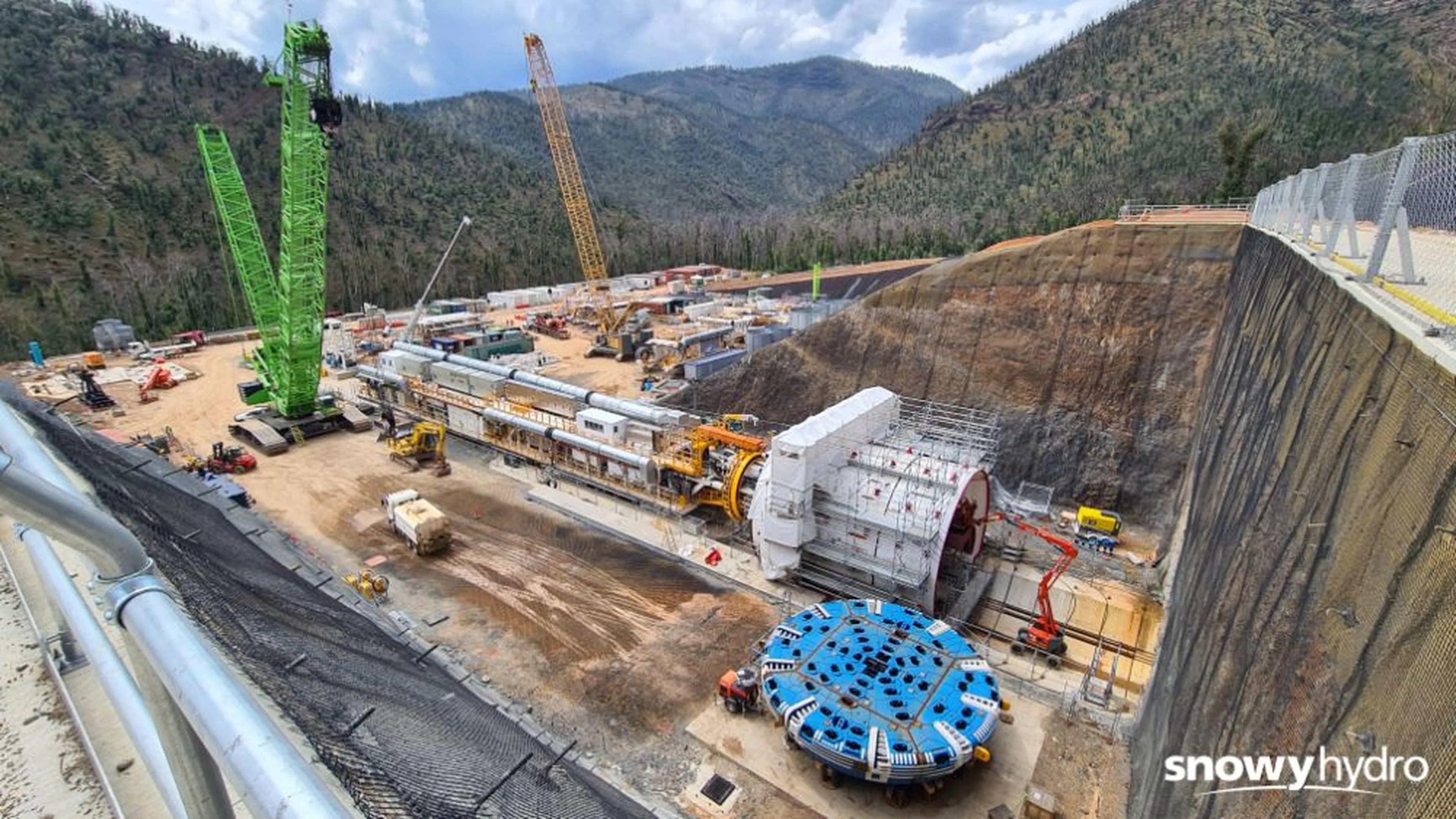Construction site with large machinery, cranes, and a tunnel-boring machine in a mountainous area under a partly cloudy sky. Snowy Hydro logo visible.