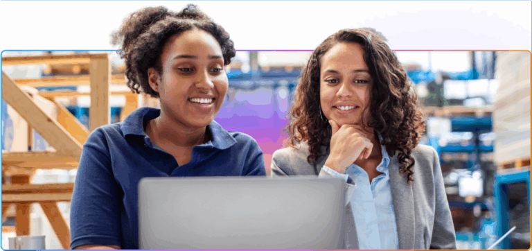Two smiling women in a warehouse setting looking at a laptop together, appearing engaged and collaborative.