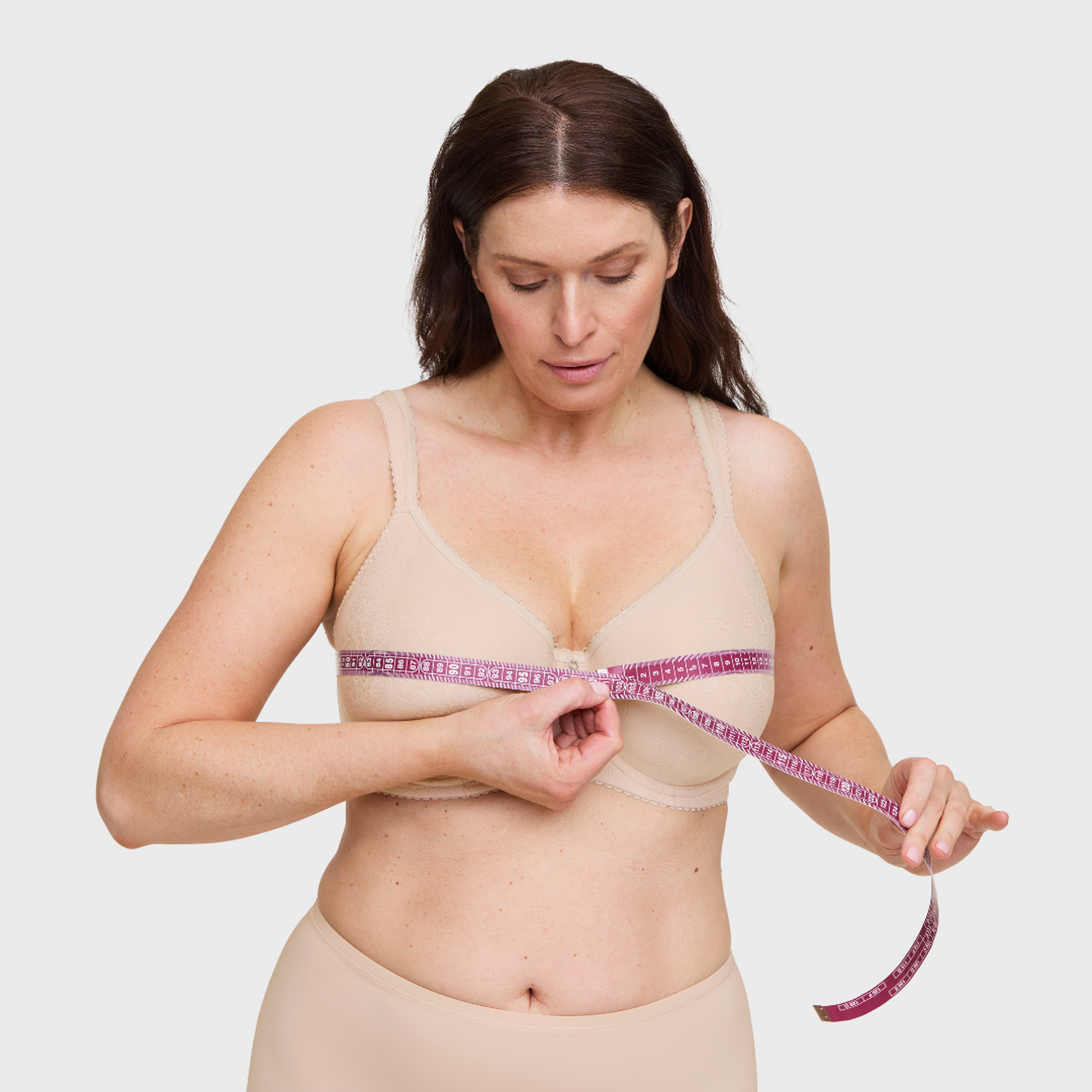 Woman in nude bra and underwear measuring her bust size with a pink measuring tape against a light gray background.