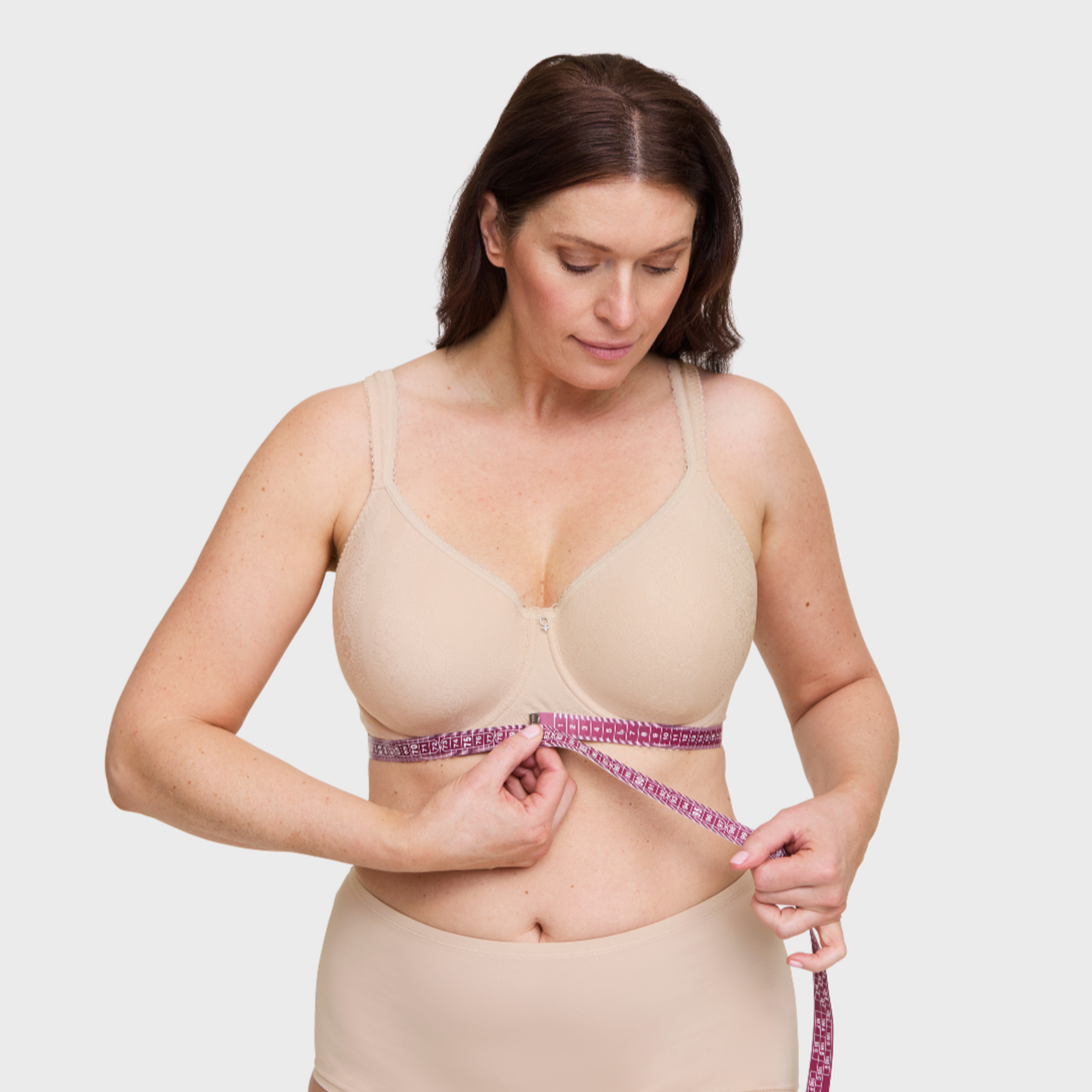 Woman in nude wireless bra measuring her underbust with a pink measuring tape for bra fitting guidance.