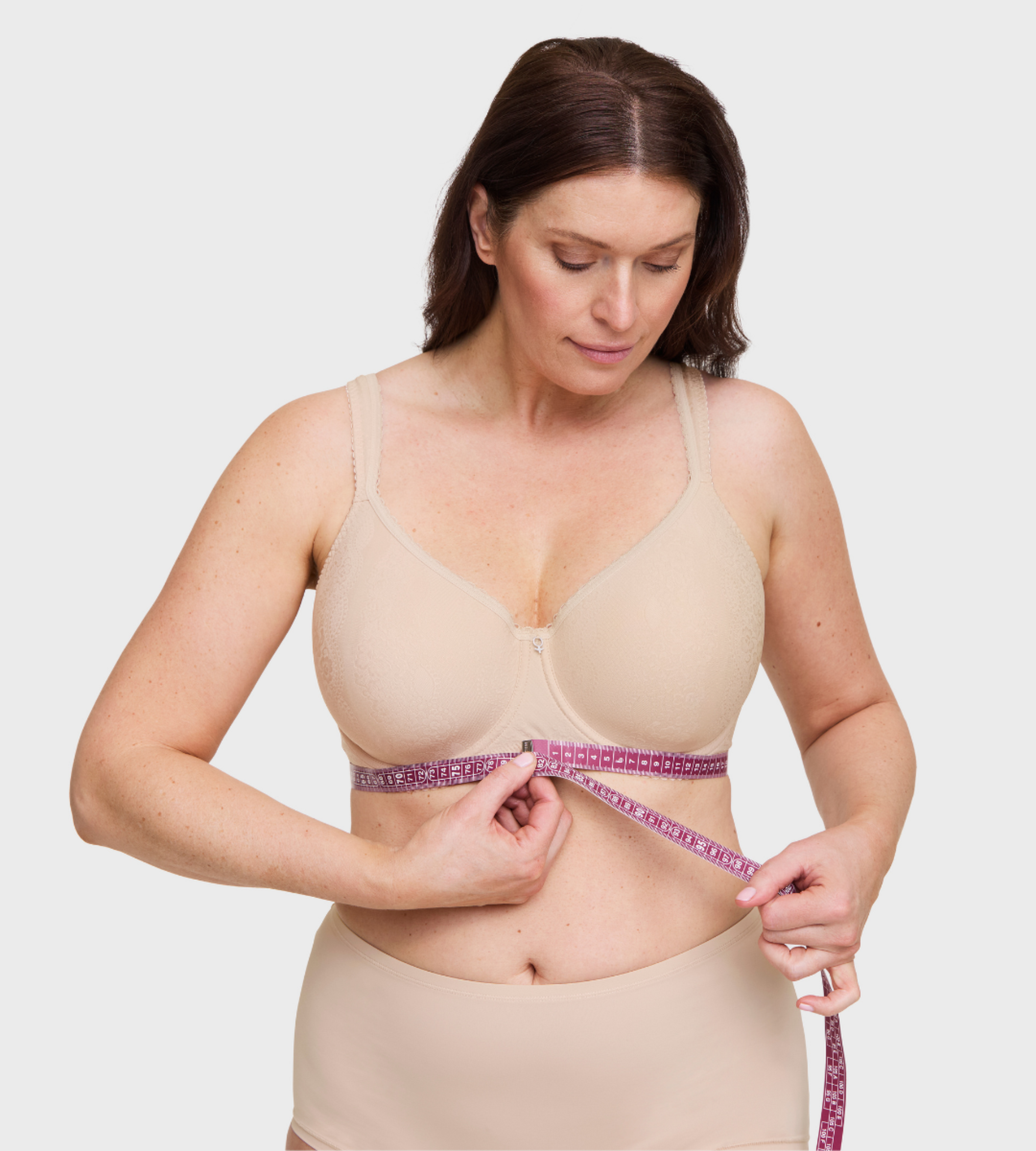 Woman in nude bra and underwear measuring her underbust with a pink tape measure for bra fitting sizing guide.