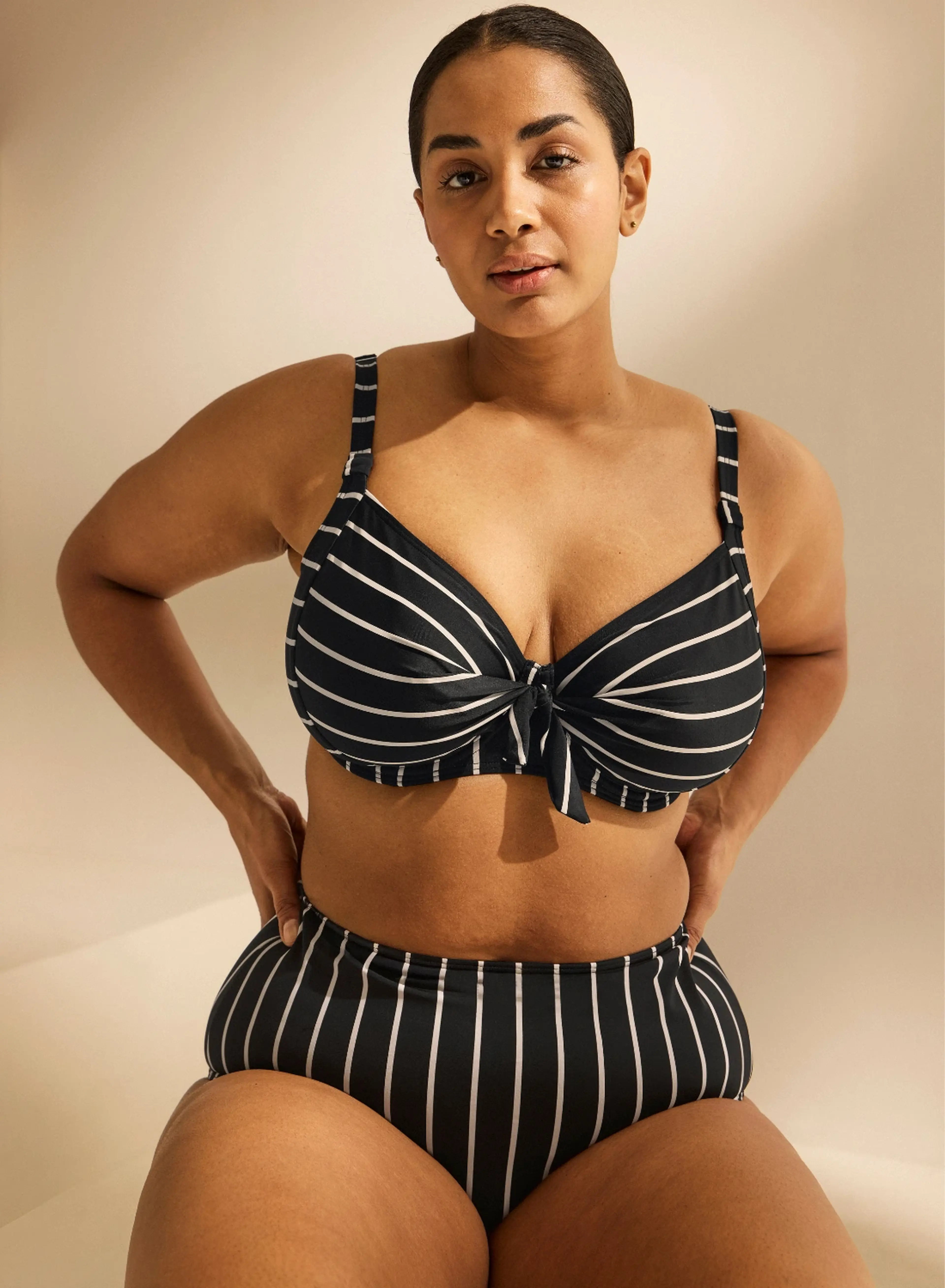 Woman in a black and white striped bikini poses confidently against a neutral background, hands on hips, with natural lighting.