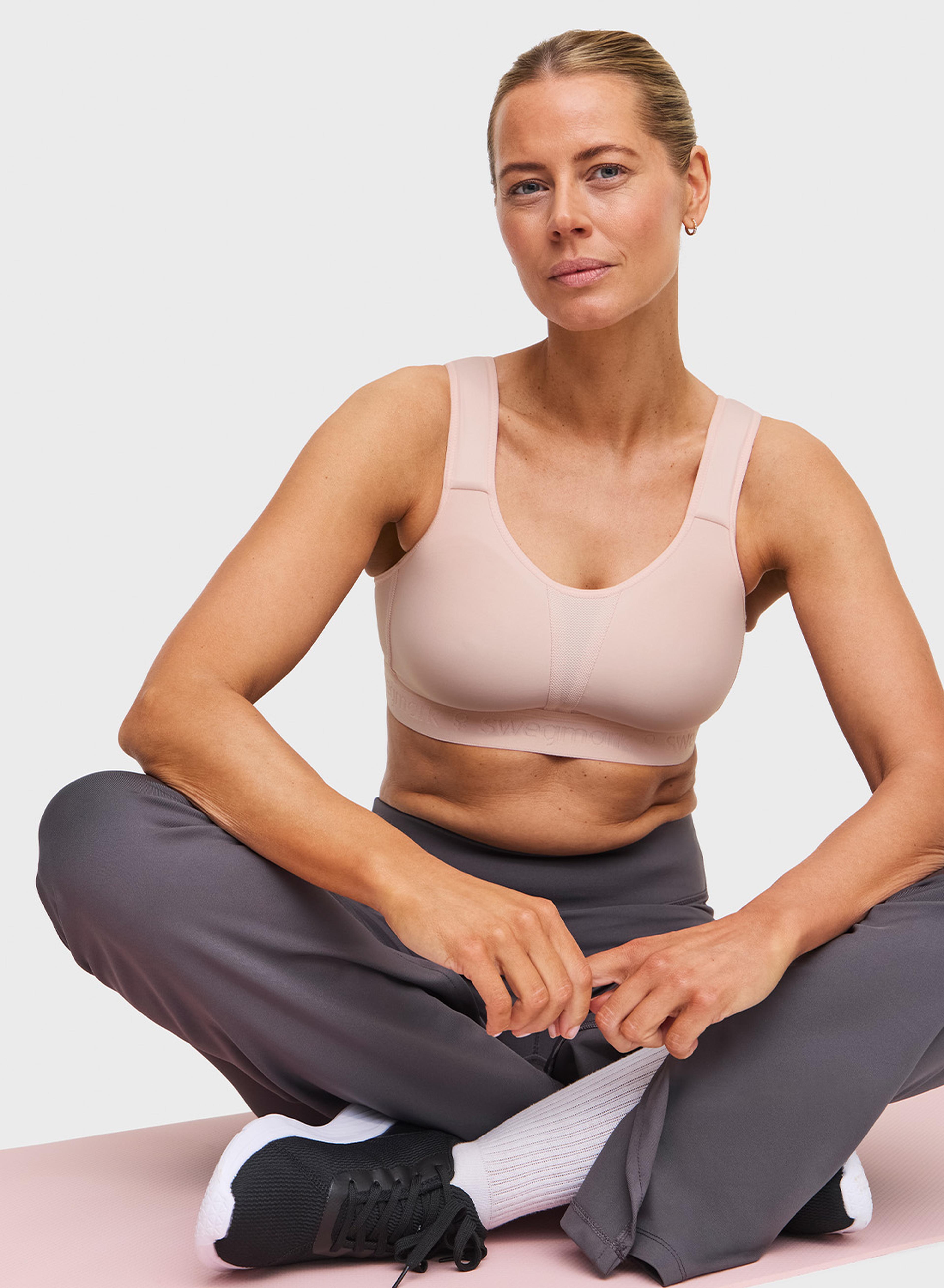 Person sitting cross-legged on a mat, wearing a pale pink sports bra, gray pants, white socks, and black shoes, against a plain background.