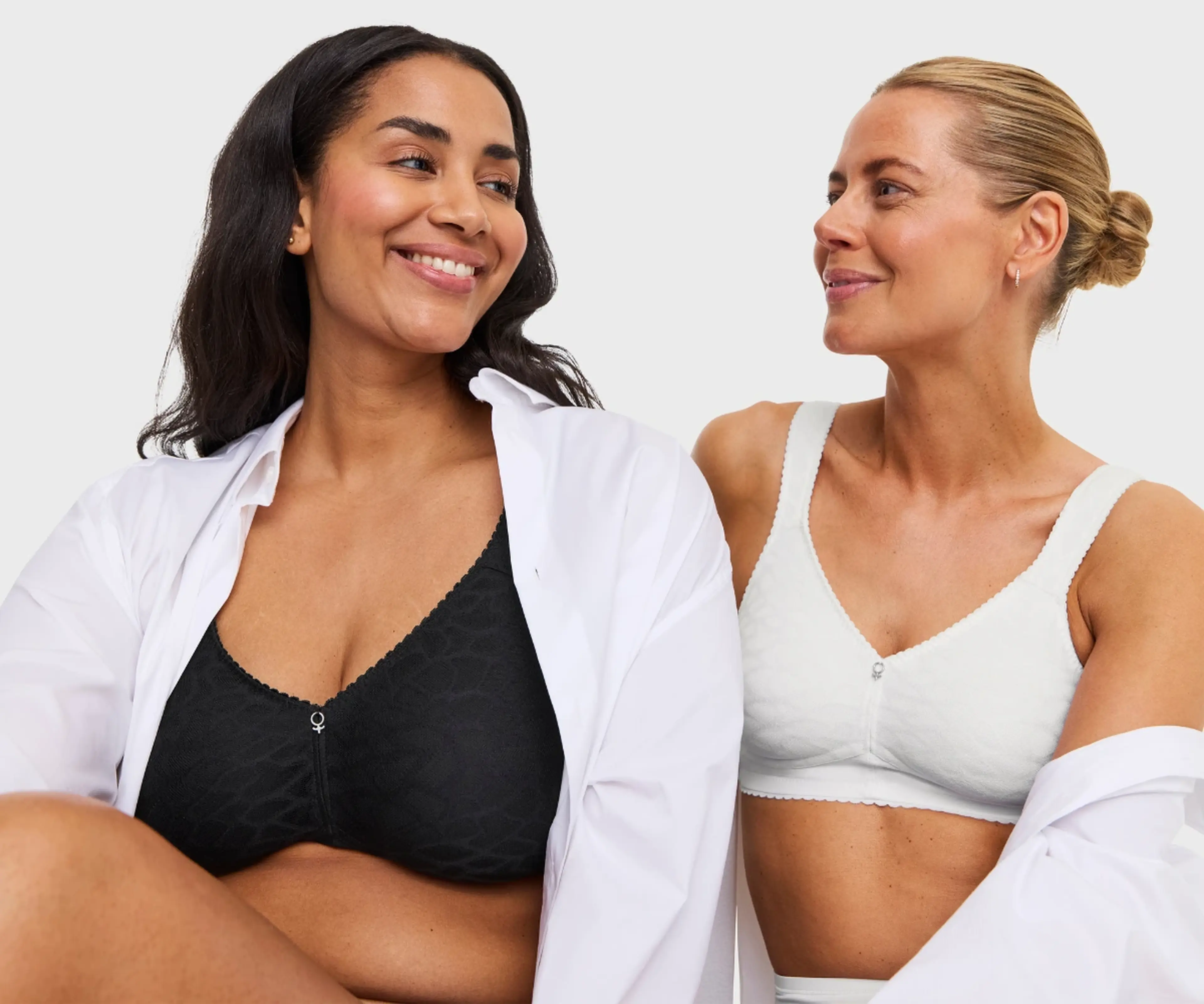 Two smiling women wearing wireless bras, one in black and one in white, with open white shirts, against a light gray background.