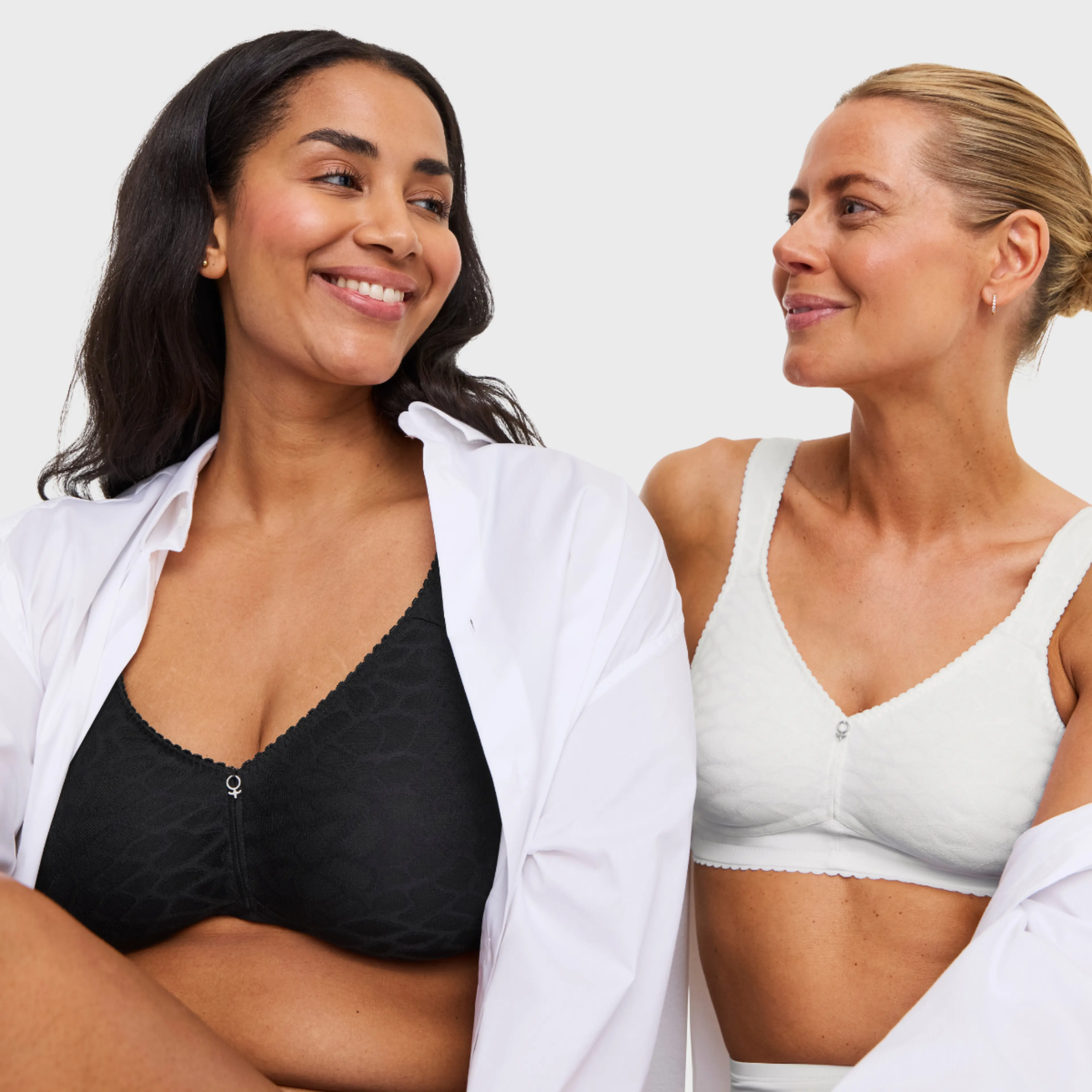 Two smiling women wearing wireless bras, one black lace and one white, with open white shirts, against a light gray background.