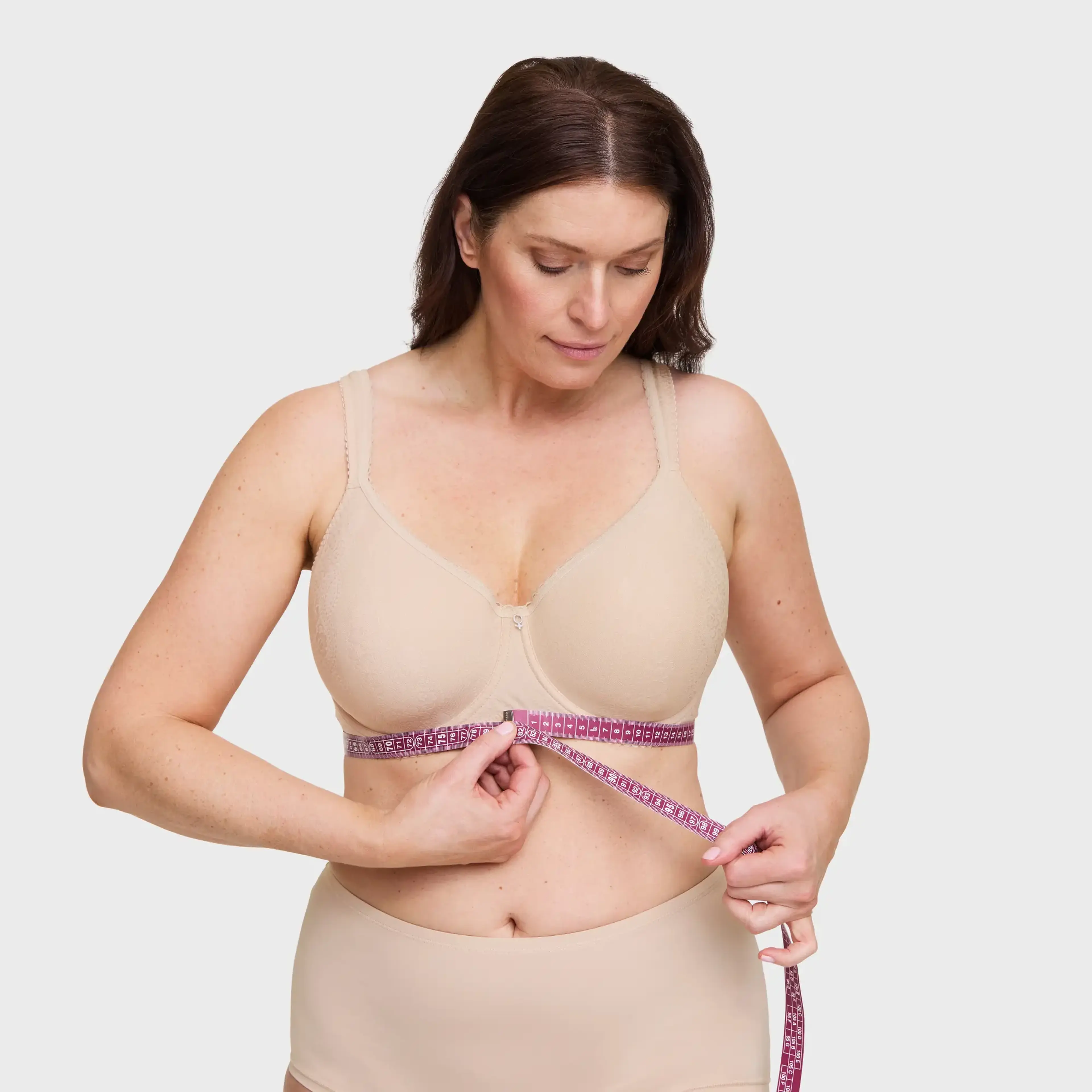Woman in nude bra and underwear measuring her underbust with a pink measuring tape against a light gray background.
