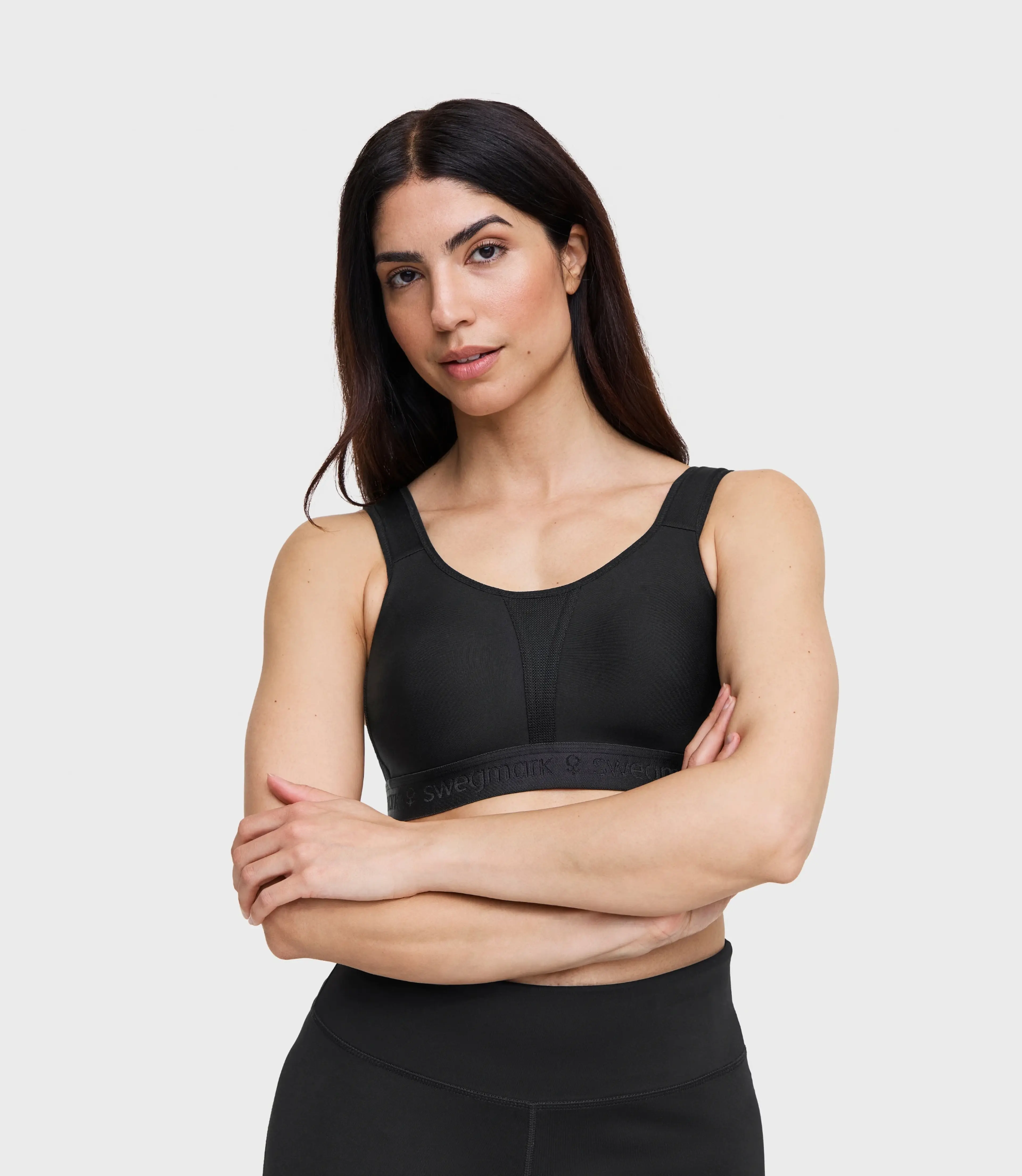 Woman with dark hair wearing a black Swegmark sports bra and matching high-waist leggings, arms crossed, on grey background.