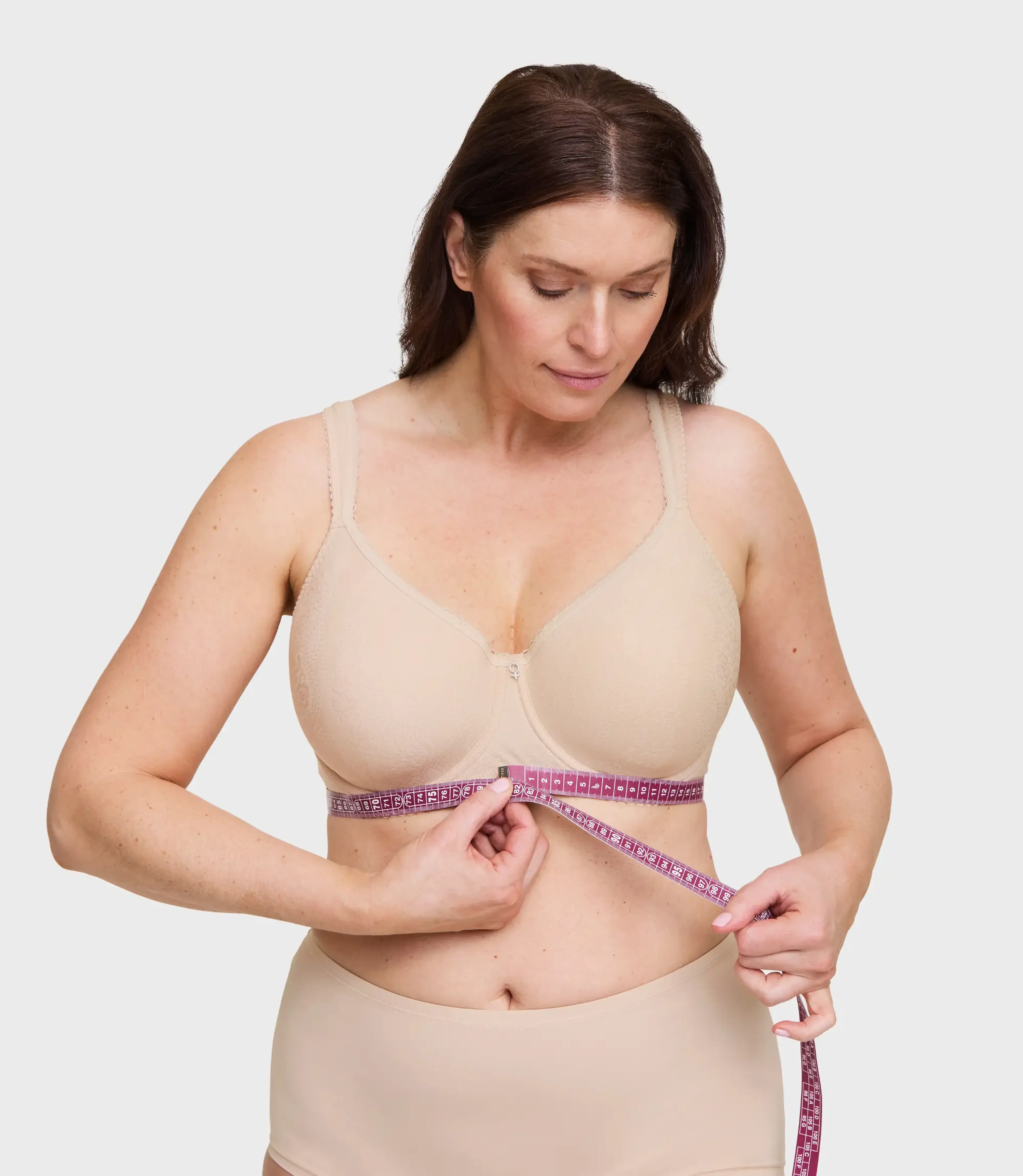 Woman in nude bra and underwear measuring her underbust with a pink tape measure against a light gray background.