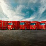 Five red trucks parked in a row on a wide, open road with a dramatic cloudy sky and distant mountains in the background.
