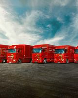 Five red trucks parked in a row on a wide, open road with a dramatic cloudy sky and distant mountains in the background.