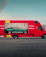 Red transport truck with a transparent side, showcasing a classic green car inside, parked in an urban area with skyscrapers in the background.