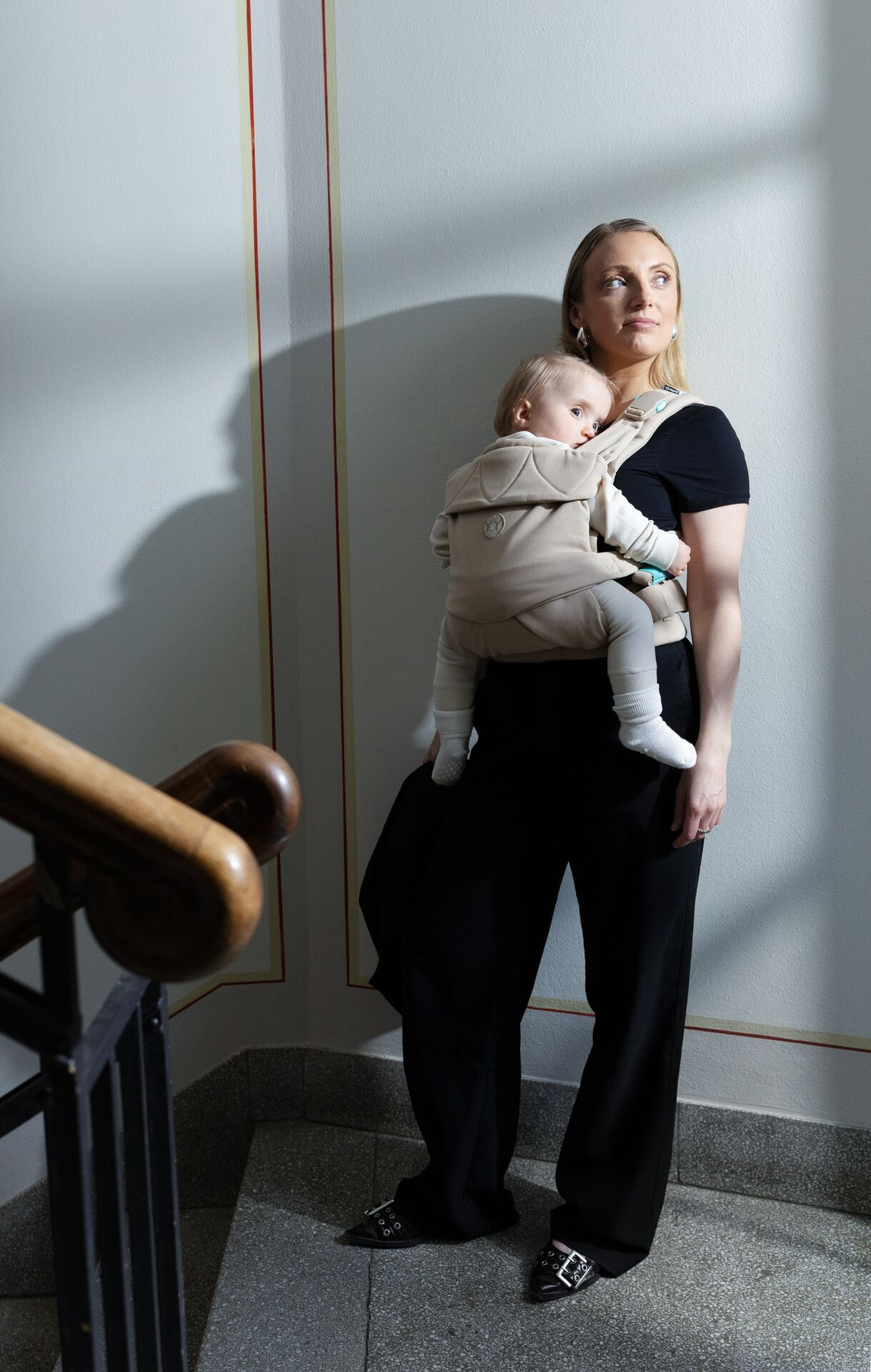 Woman in black outfit leaning against a wall, carrying a baby in a honey beige Najell Easy vol. 2 baby carrier. Sunlight casts shadows, staircase railing visible.