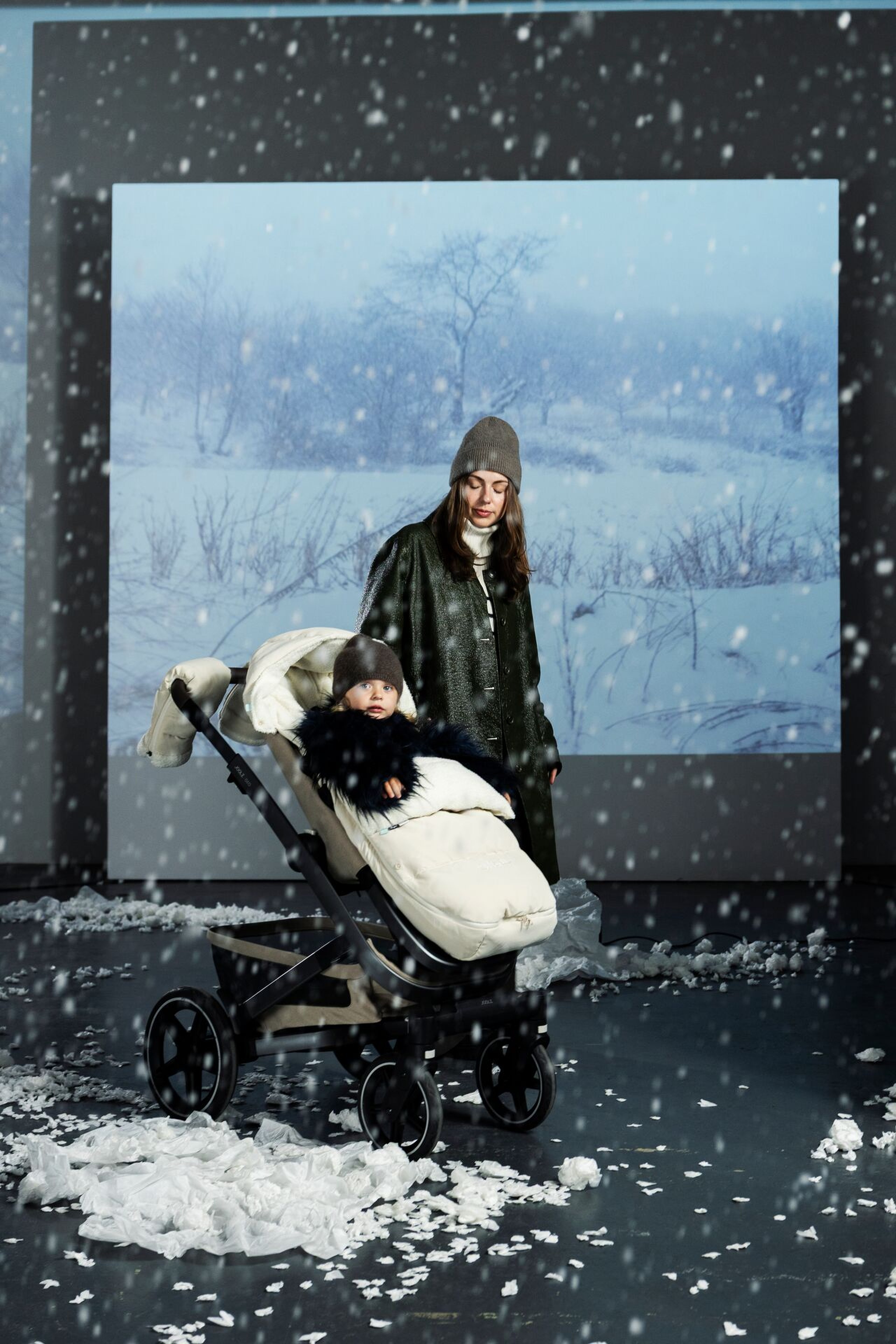 Baby in a beige footmuff with matching stroller gloves on a stroller in snowy winter weather.