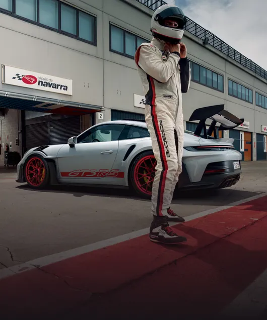 Porsche 911 GT3 RS en Gris Glacé métallisé, devant un garage. Vue de côté. Au premier plan, un pilote de course.