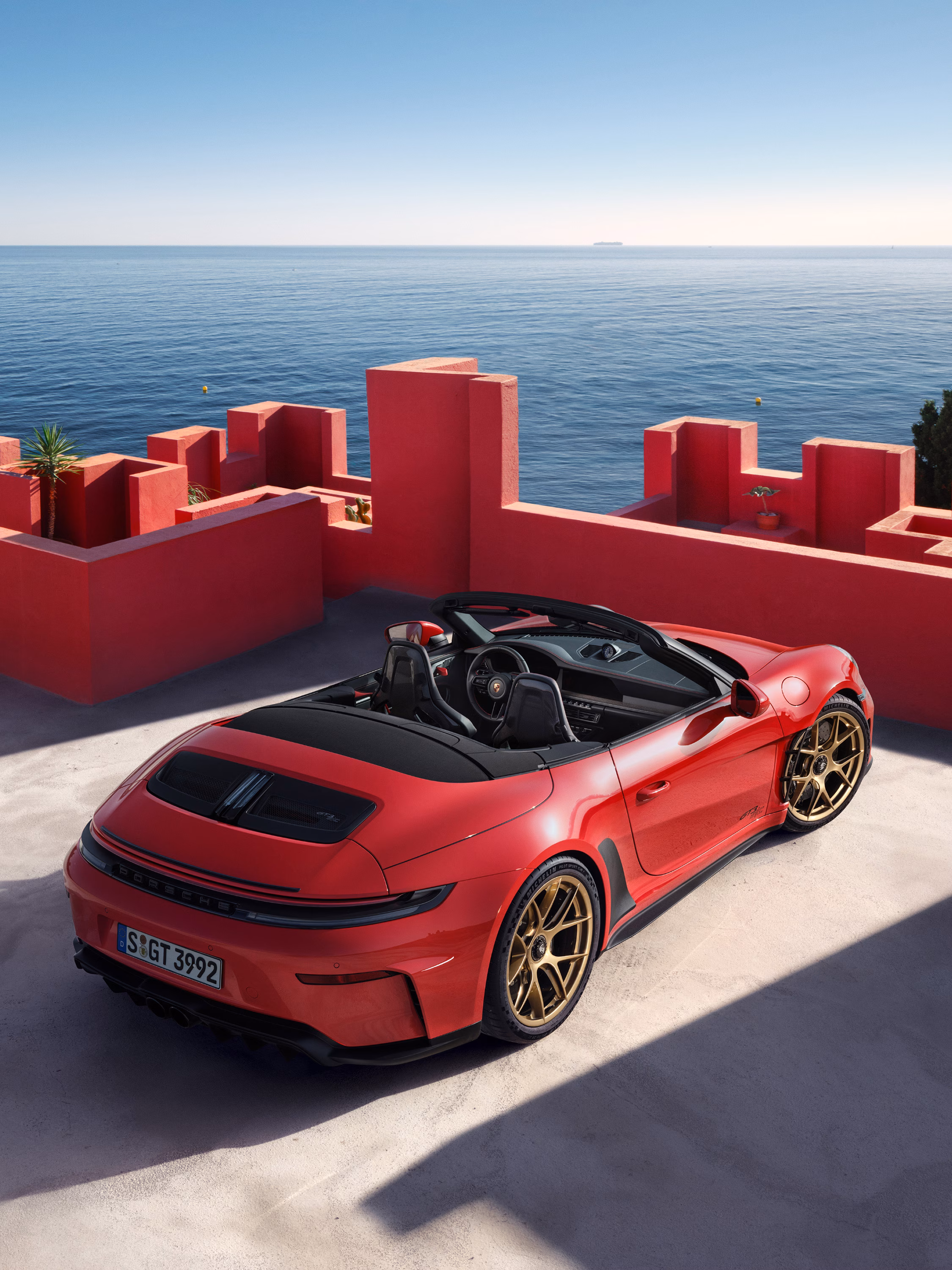 Vista trasera y lateral de un Porsche 911 GT3 S/C en color Rojo Guardia delante de bloques de hormigón de diferentes tonalidades de color rojizo. Al fondo se ve el mar.