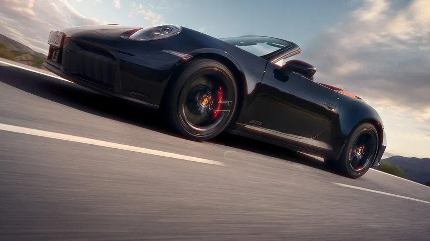 The new 911 Carrera GTS Cabriolet, in Jet Black Metallic, driving across a road, clouds can be seen in the sky.