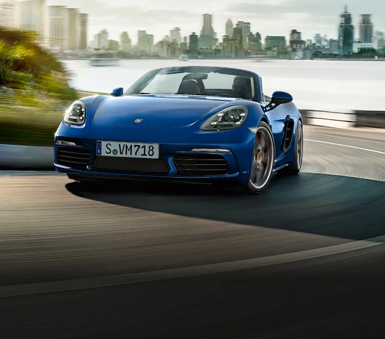 海岸沿いの道路でカーブを曲がるゲンチアンブルー メタリックの718 Boxster S。フロントビューとサイドビュー