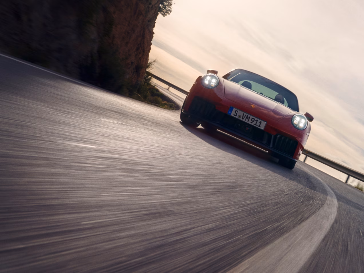 Porsche 911 Targa 4 GTS in Carmine Red driving through a corner on a road in a mountainous landscape. Front view from the front.