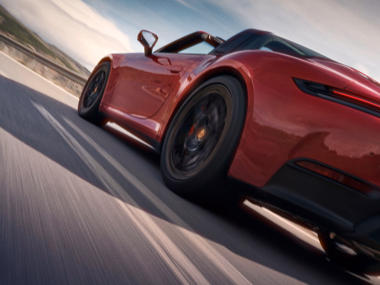 Porsche 911 Targa 4 GTS in Carmine Red driving on a road through a mountainous landscape. View of the side and rear end from behind.