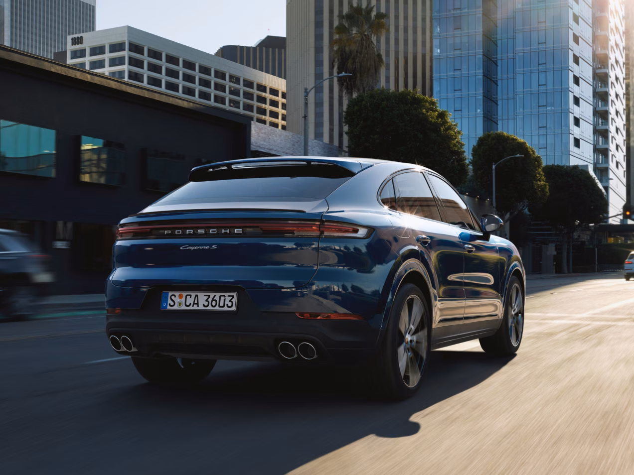 Porsche Cayenne S Coupé in Algarve Blue driving through a city. Rear and side view from behind.