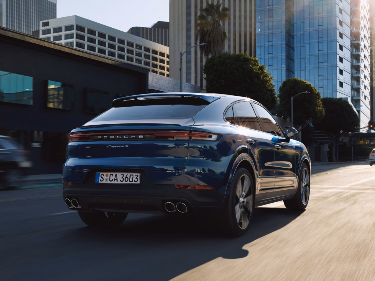 Porsche Cayenne S Coupé in Algarve Blue driving through a city. Rear and side view from behind.