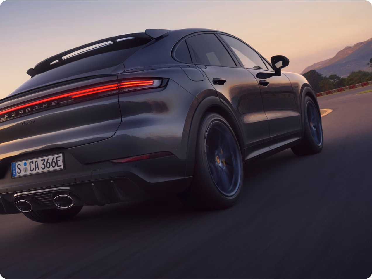 Un Porsche Cayenne Turbo E-Hybrid Coupé con paquete GT en color Gris Cuarcita Metalizado rueda en un circuito. Vista desde abajo de la parte trasera y el lateral.