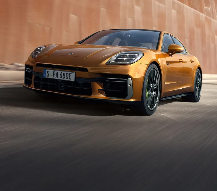 Porsche Panamera Turbo E-Hybrid i Madeira Gold Metallic kör förbi en vägg av metall. Fronten och sidan sett framifrån.