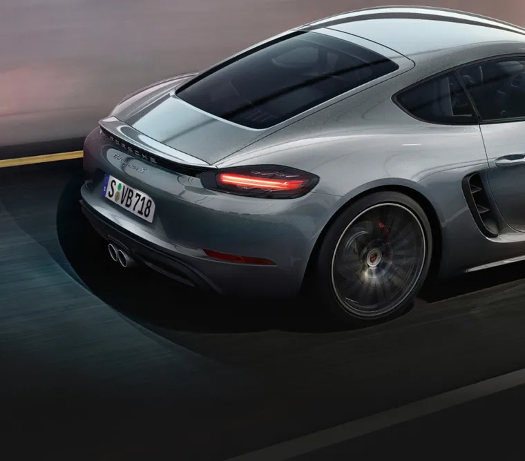 Porsche 718 Cayman S цвета серебристый металлик (GT Silver Metallic) едет по дороге. Вид на заднюю часть кузова с задним антикрылом.