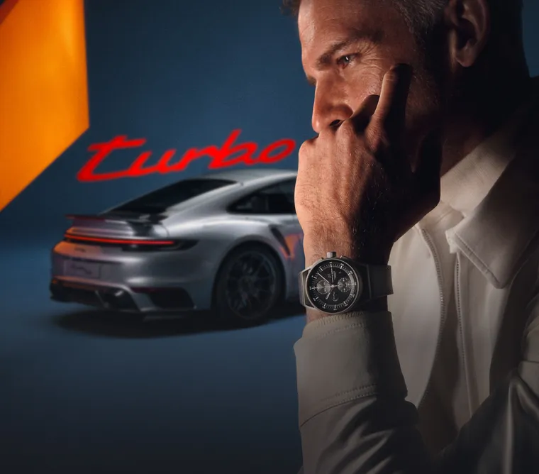 A man with the Porsche Design Chronograph 911 Turbo 50 Years on his wrist is shown in front of a Porsche 911 Turbo 50 Years in GT Silver Metallic. 