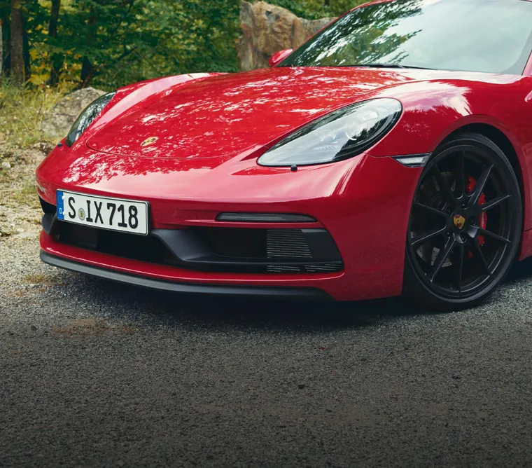 Porsche 718 Cayman GTS 4.0 in Rosso Carminio parcheggiata sul ciglio della strada nella foresta. Vista anteriore con frontale specifico della GTS 4.0.