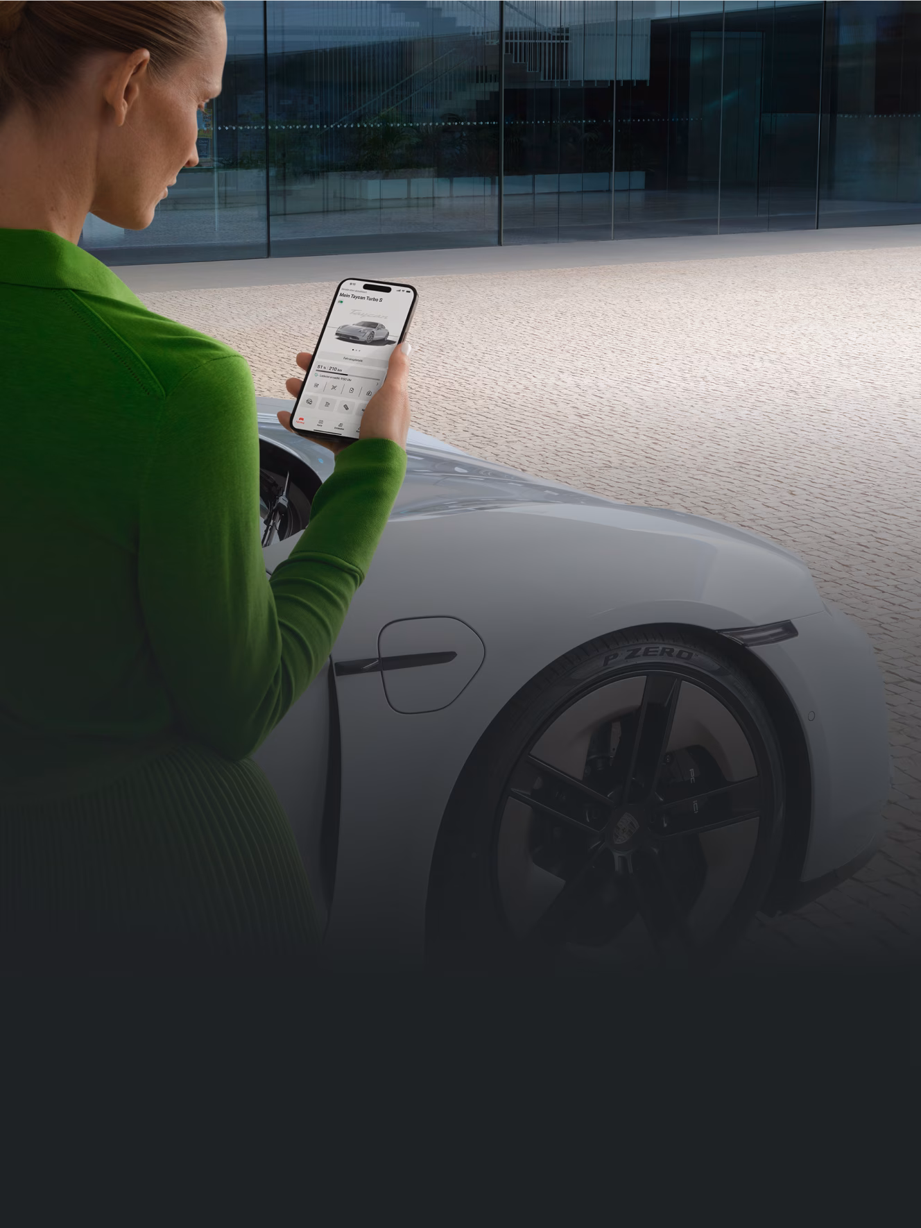A woman looks at her smartphone and uses Porsche Connect. A Porsche Taycan Turbo S is parked in the background.
