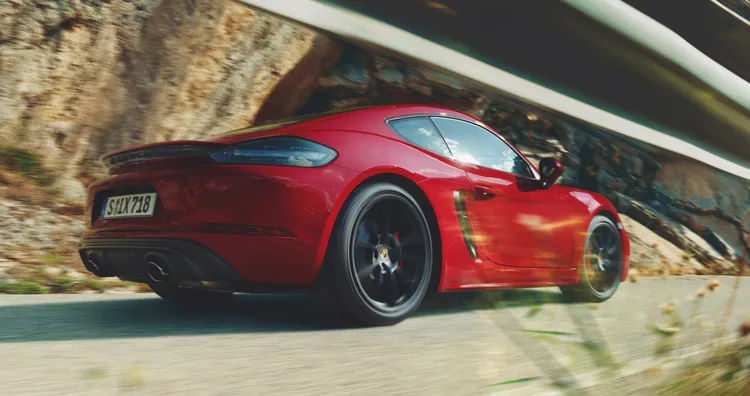 718 Cayman GTS 4.0 en color Rojo Carmín circulando por una carretera costera. Vista de la parte trasera y el lateral desde una perspectiva baja.