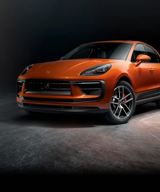 Un Porsche Macan S en color Papaya Metalizado frente a un fondo oscuro. Vista frontal y lateral.