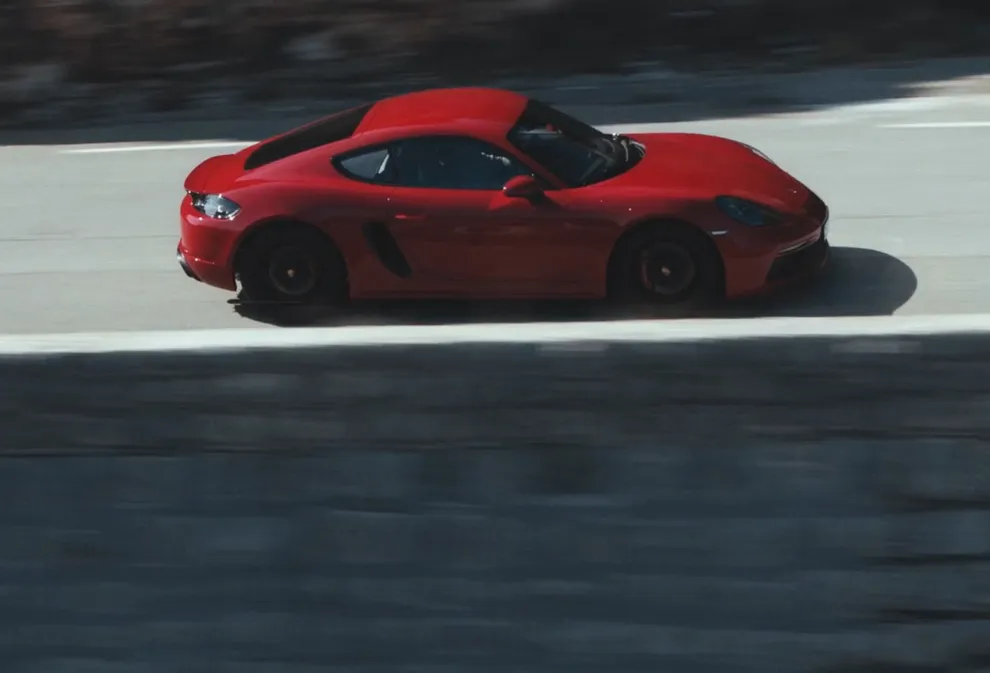 Porsche 718 Cayman GTS 4.0 in Carmine Red driving along a mountain road. Side view.